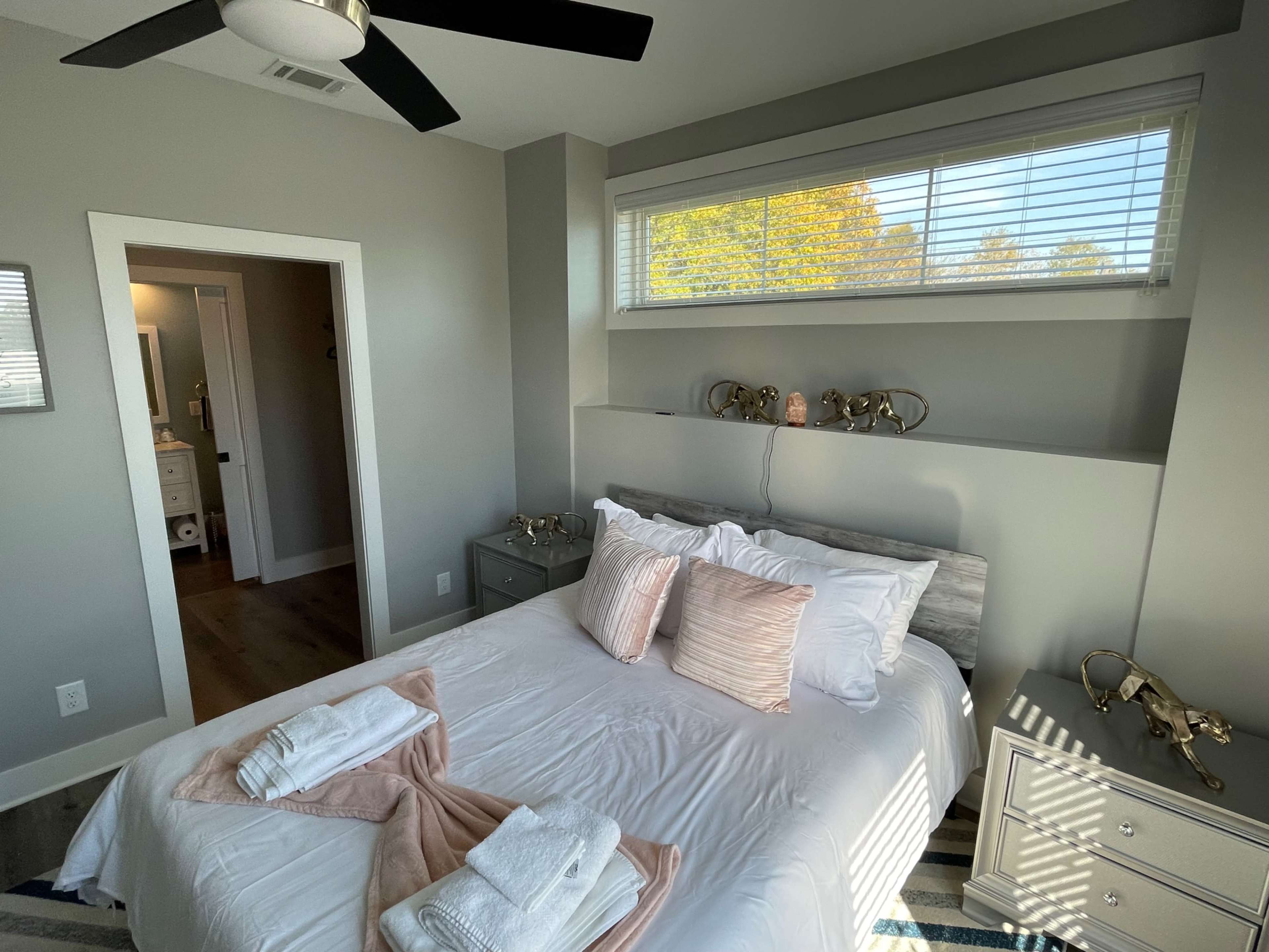 The image features a neatly arranged bedroom with a queen-sized bed adorned with pillows and a folded blanket, a window with blinds allowing natural light, and two nightstands with decorative lamps on either side.