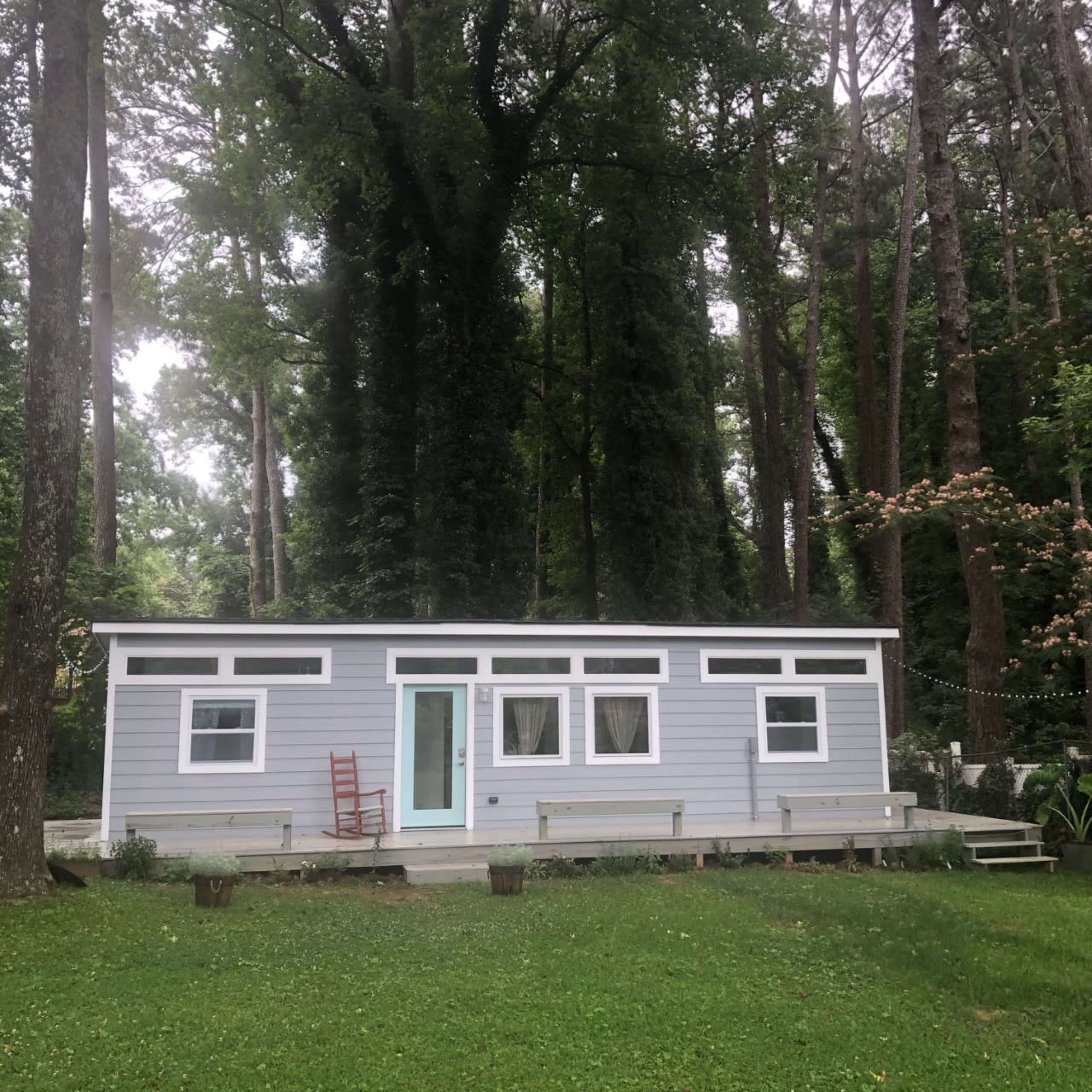 A small, light gray house with a turquoise door and a red rocking chair is surrounded by tall trees and green grass.
