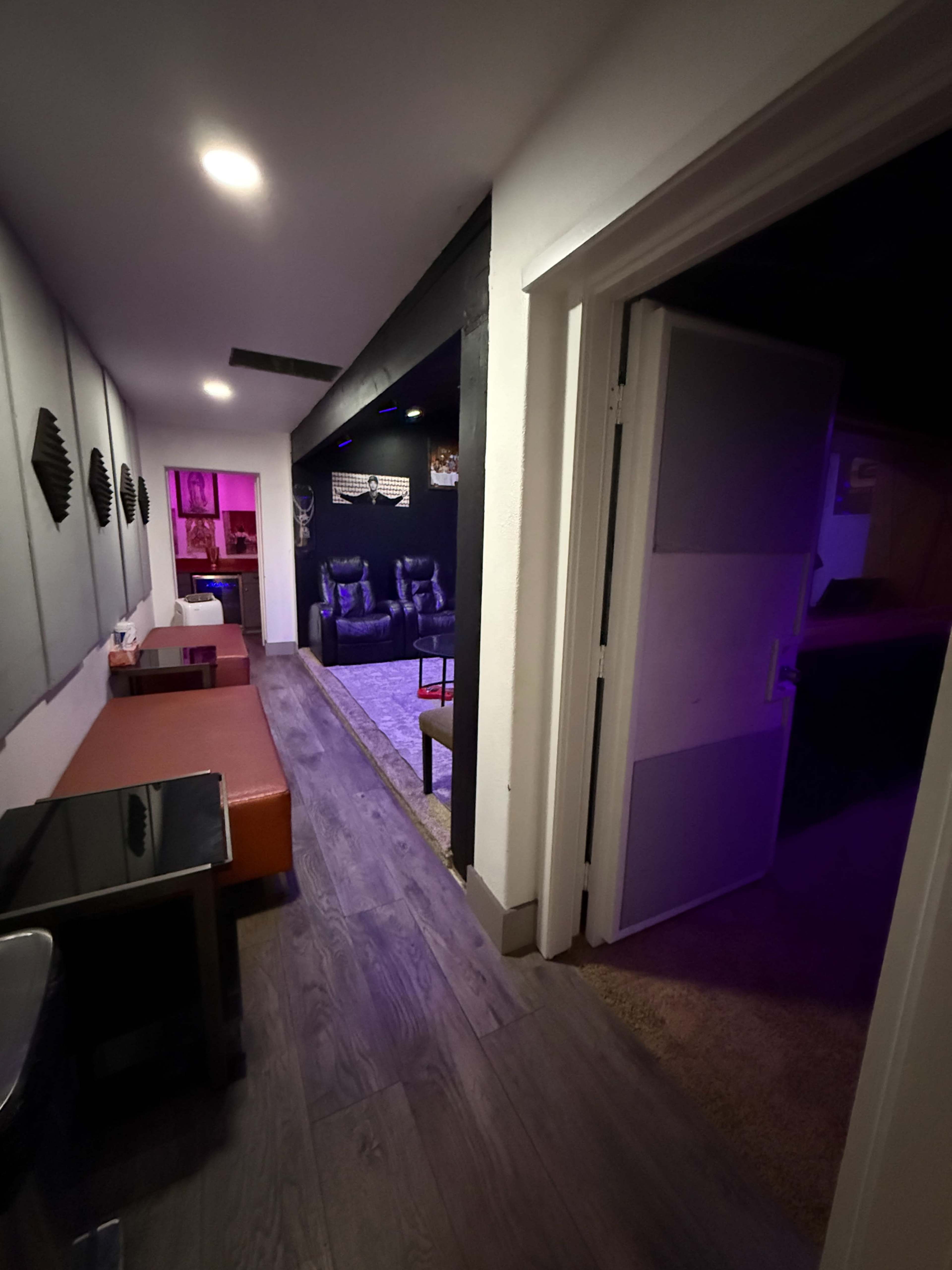 The image shows a dimly lit hallway leading to a room with seating and colorful lighting in the background.