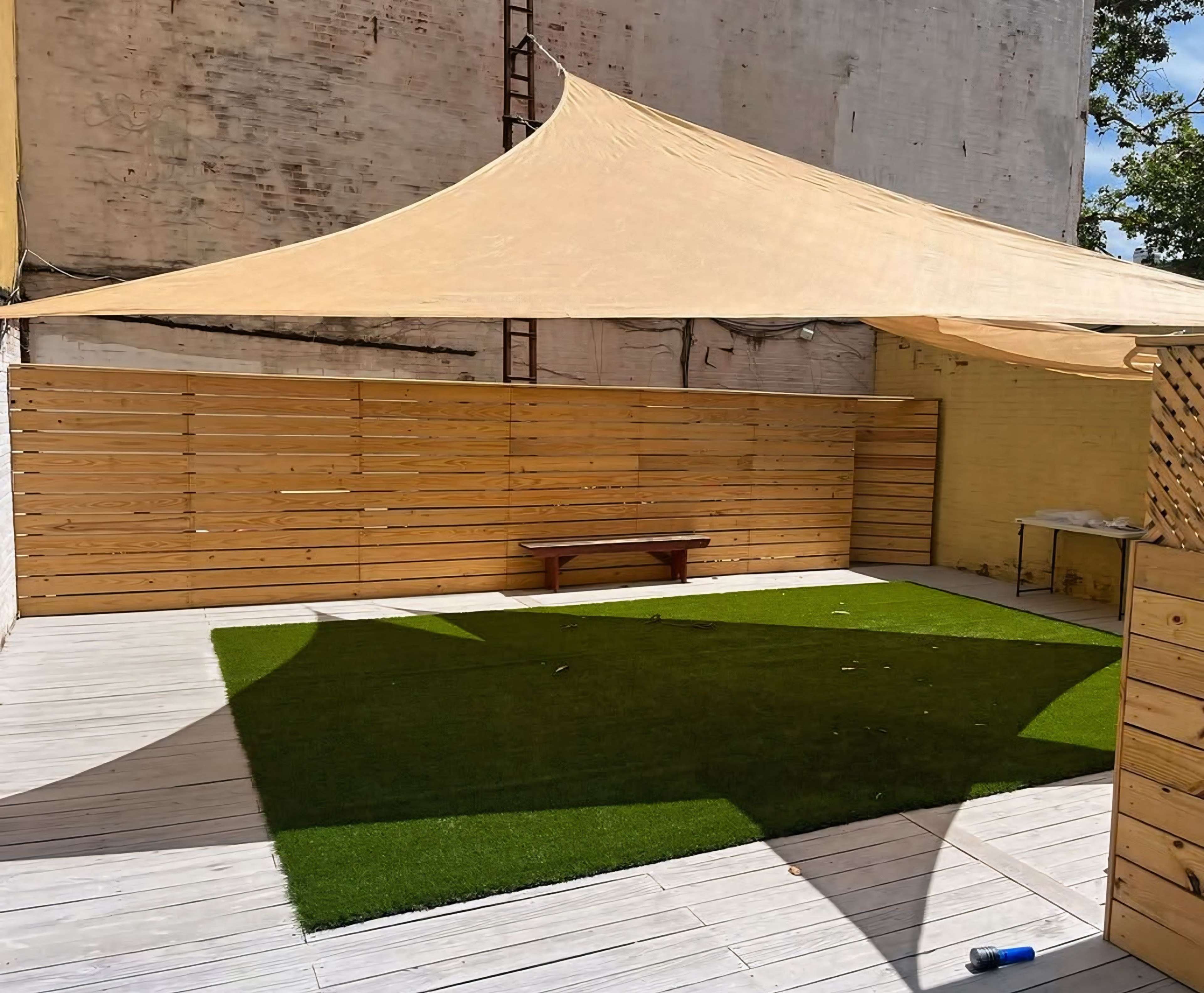 The image shows a courtyard with a wooden fence, a large tan fabric canopy overhead, a small bench, and a patch of artificial grass on a wooden deck.