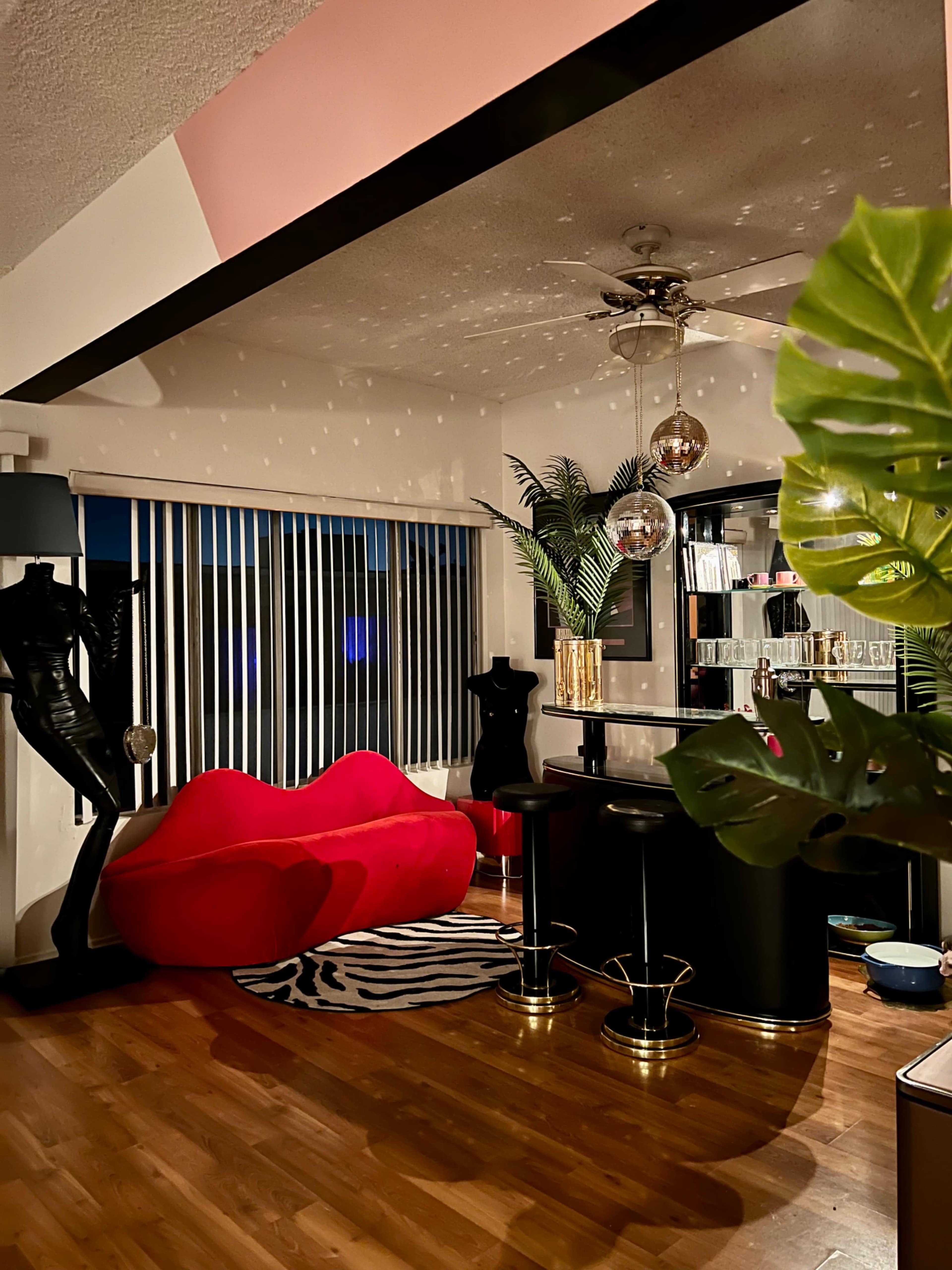 A stylish living area featuring a red couch, a black bar with stools, and decorative plants, illuminated by a ceiling fan and pendant lights.