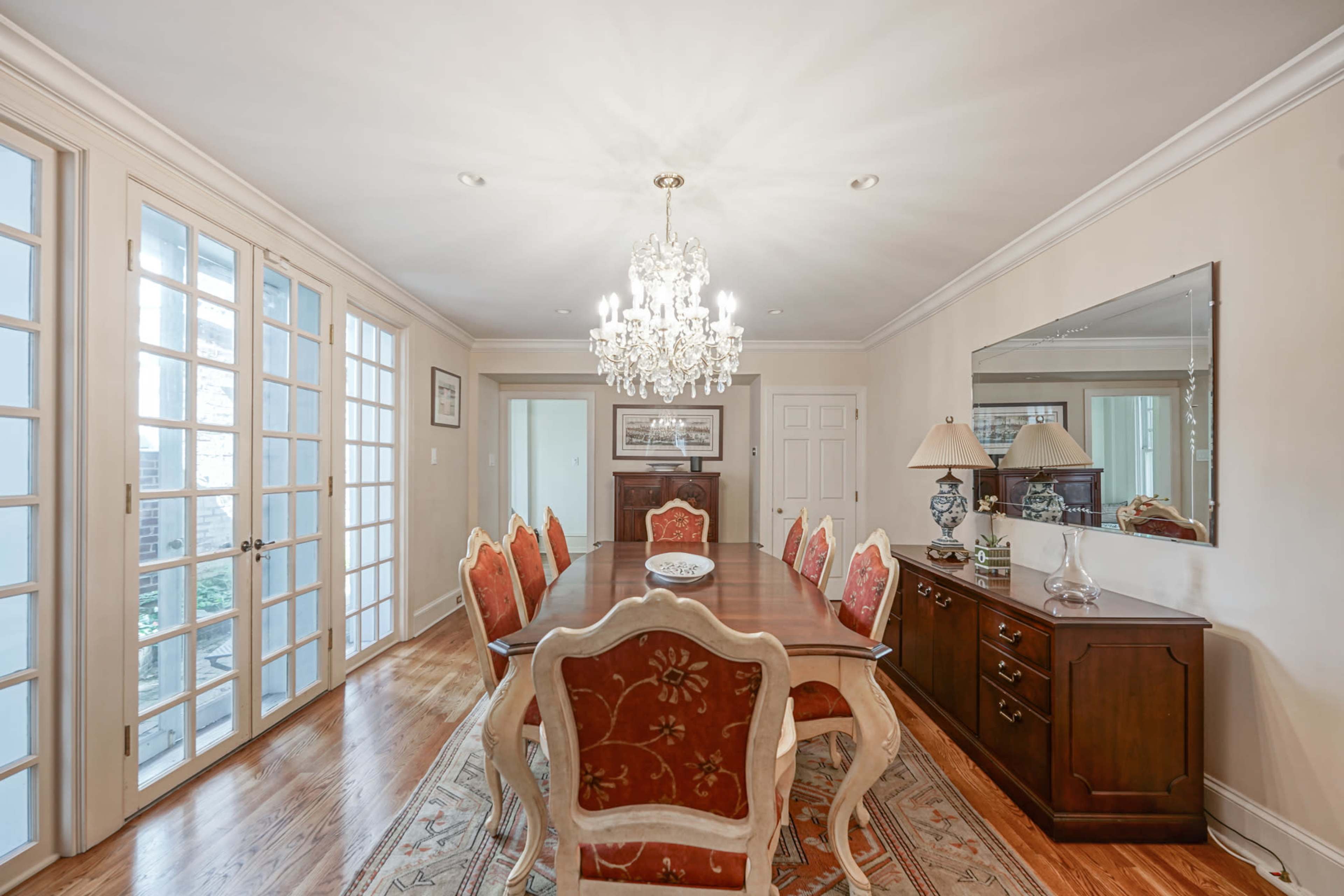 A formal dining room features a long table surrounded by ornate chairs, a chandelier overhead, and glass doors leading to an outdoor area.