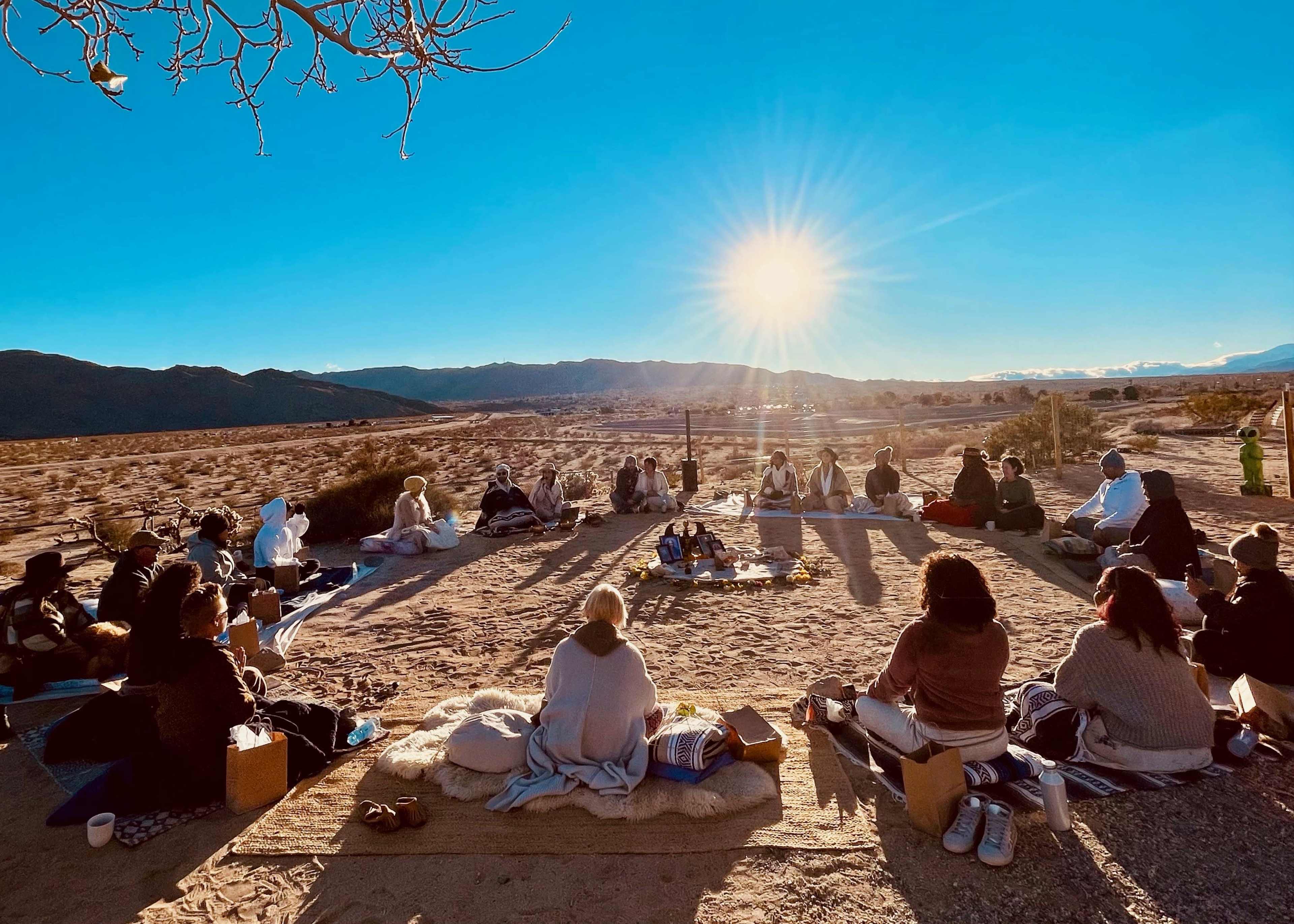 A group of people sits in a circle on the ground during a gathering at sunset in a desert landscape.