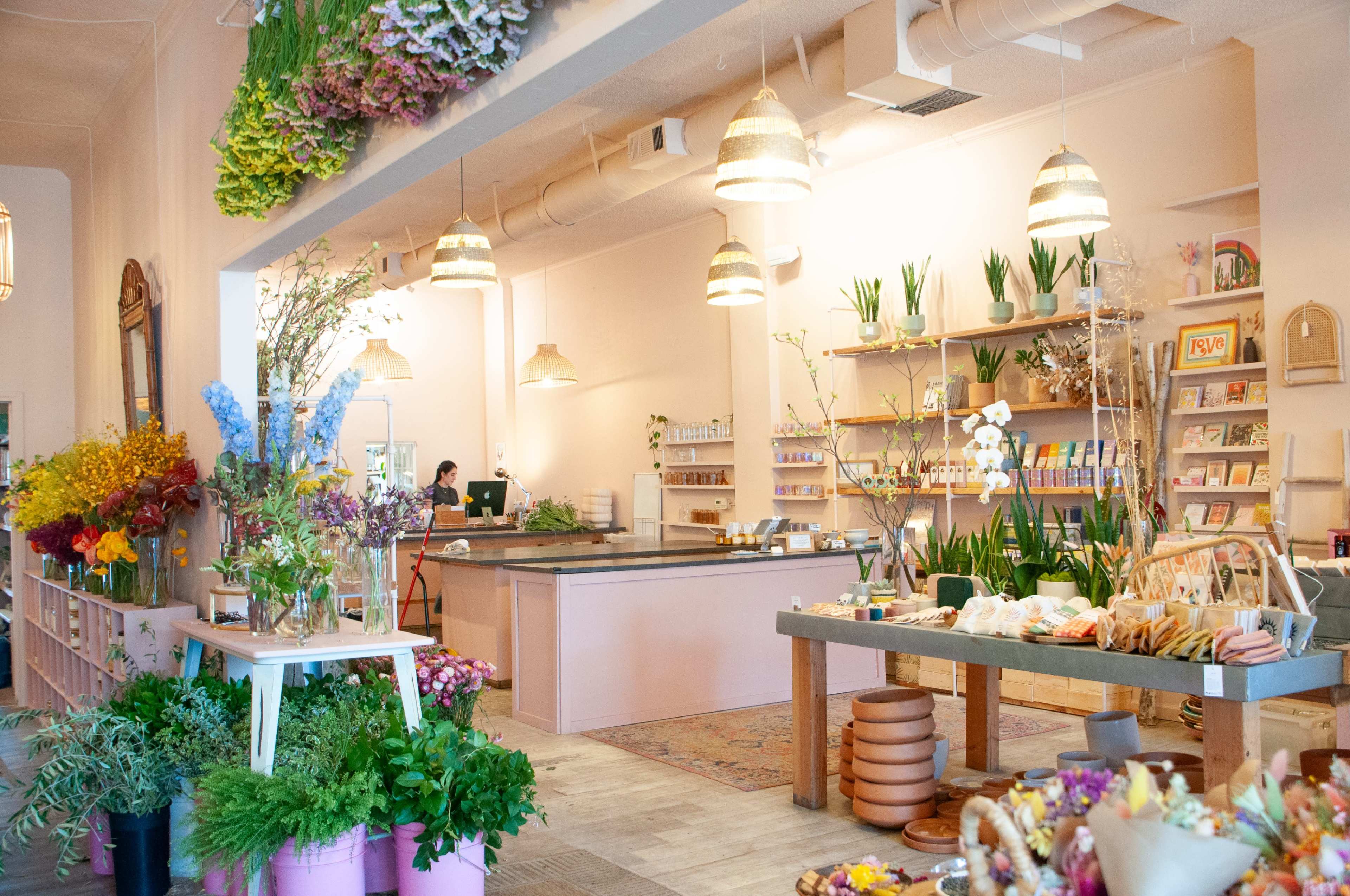 A bright floral shop features an open layout with shelves stocked with plants and flowers, a central countertop, and pendant lighting.