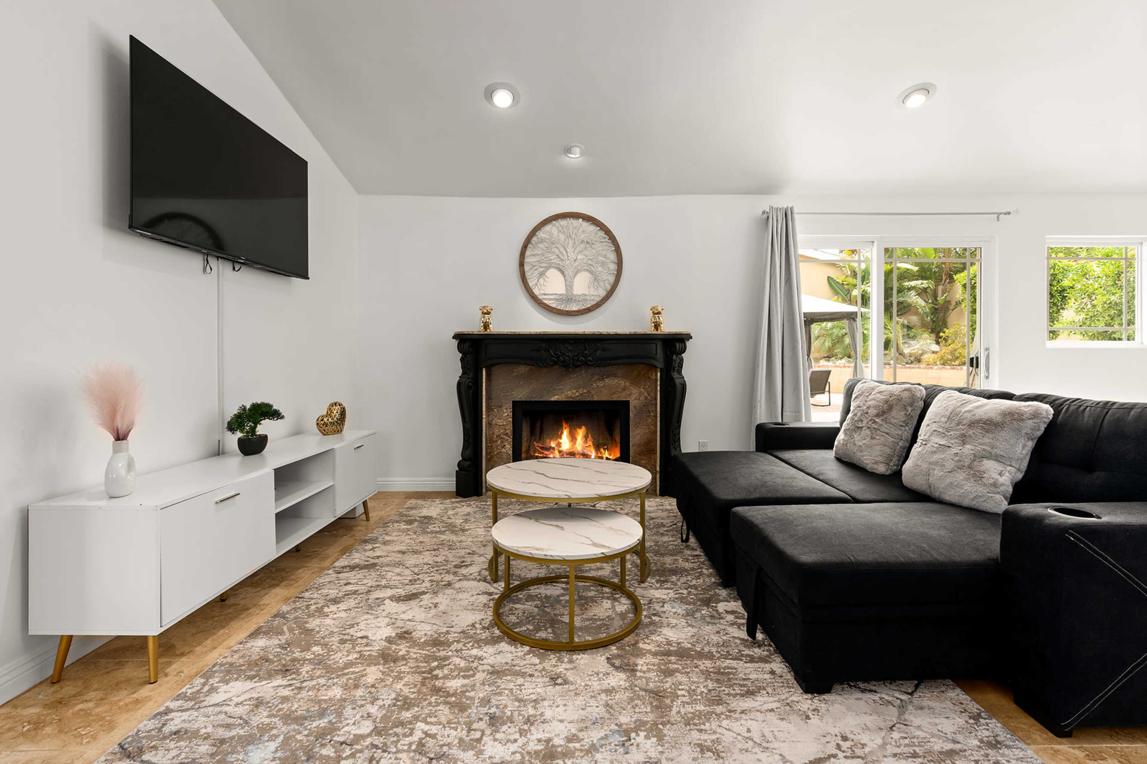 A modern living room features a black sectional sofa, a marble-top coffee table, a fireplace with a decorative mantel, and a wall-mounted television.