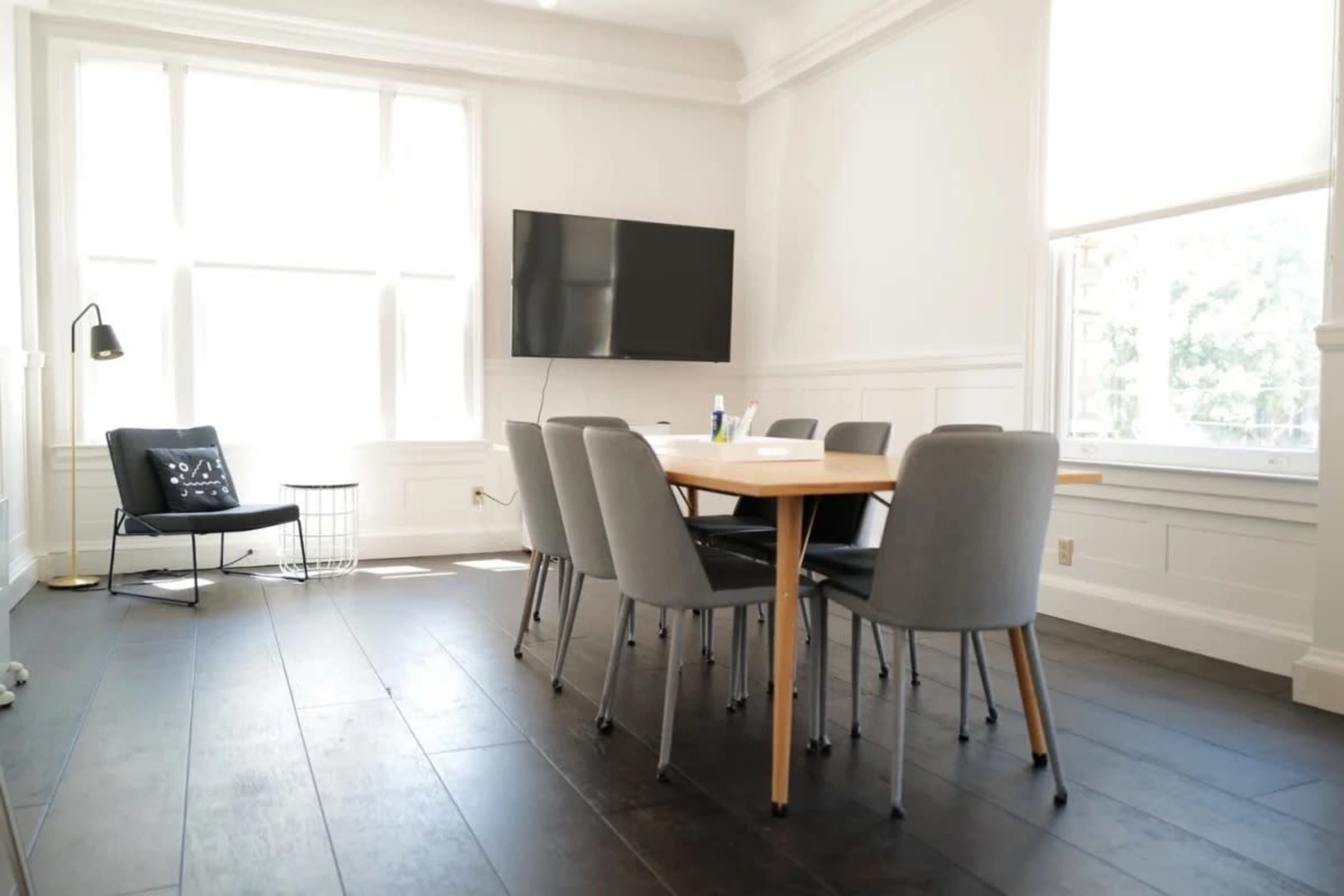 A modern meeting room features a wooden table surrounded by six gray chairs, with a television mounted on the wall and large windows allowing natural light.