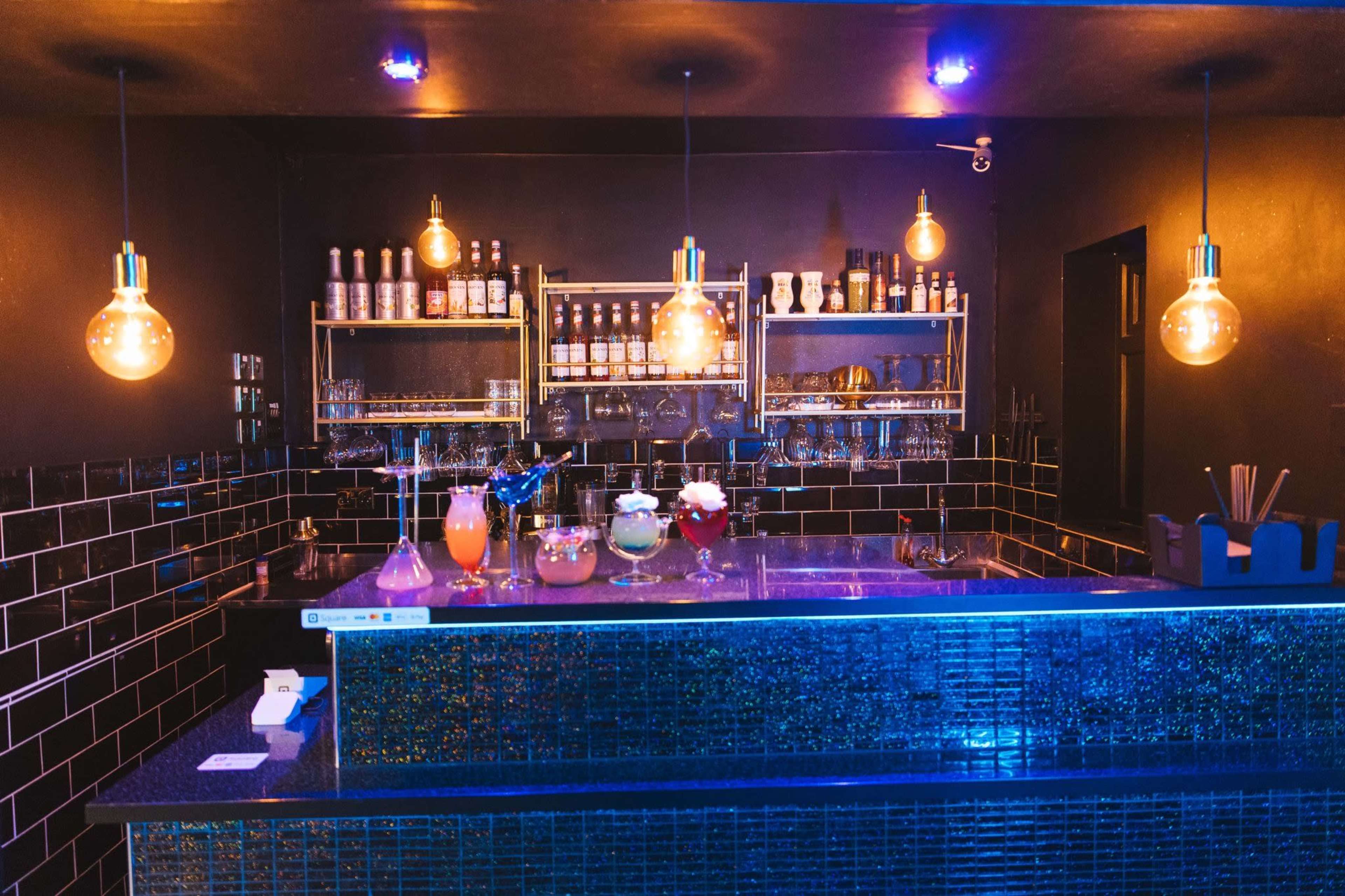 The image shows a stylish bar with a variety of colorful cocktails displayed on the counter and bottles arranged on shelves behind.