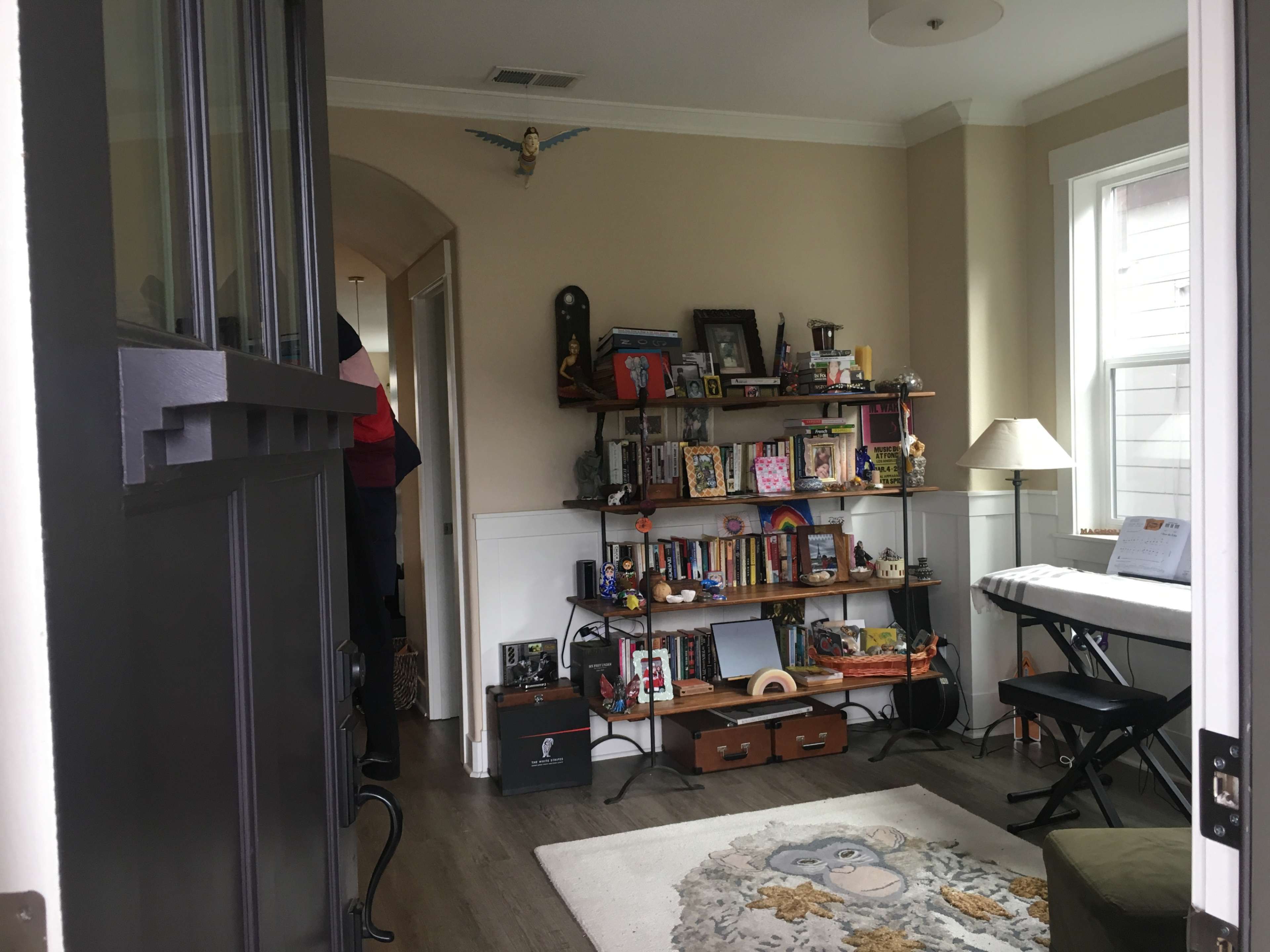A view of a cozy living area from an open door, featuring a bookshelf filled with various items and a keyboard near a window.