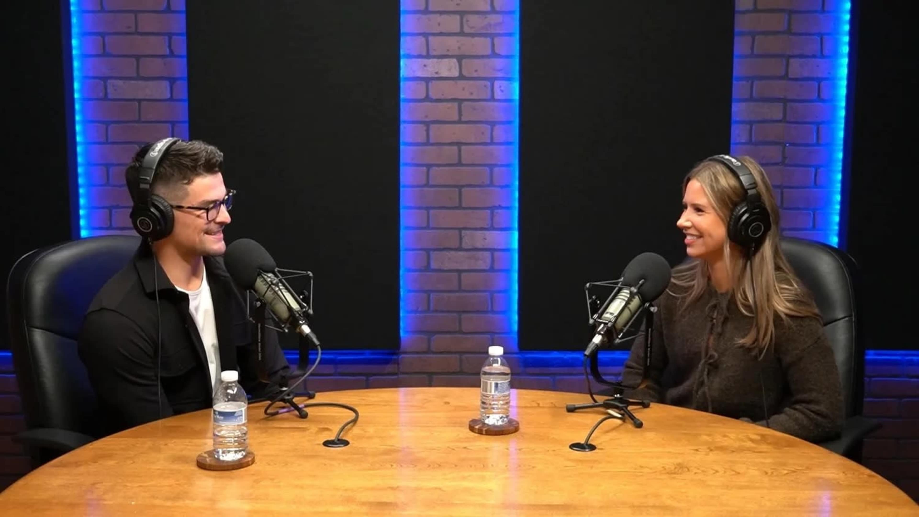 Two individuals are seated at a wooden table in a studio, each wearing headphones and speaking into microphones, with blue lighting in the background.