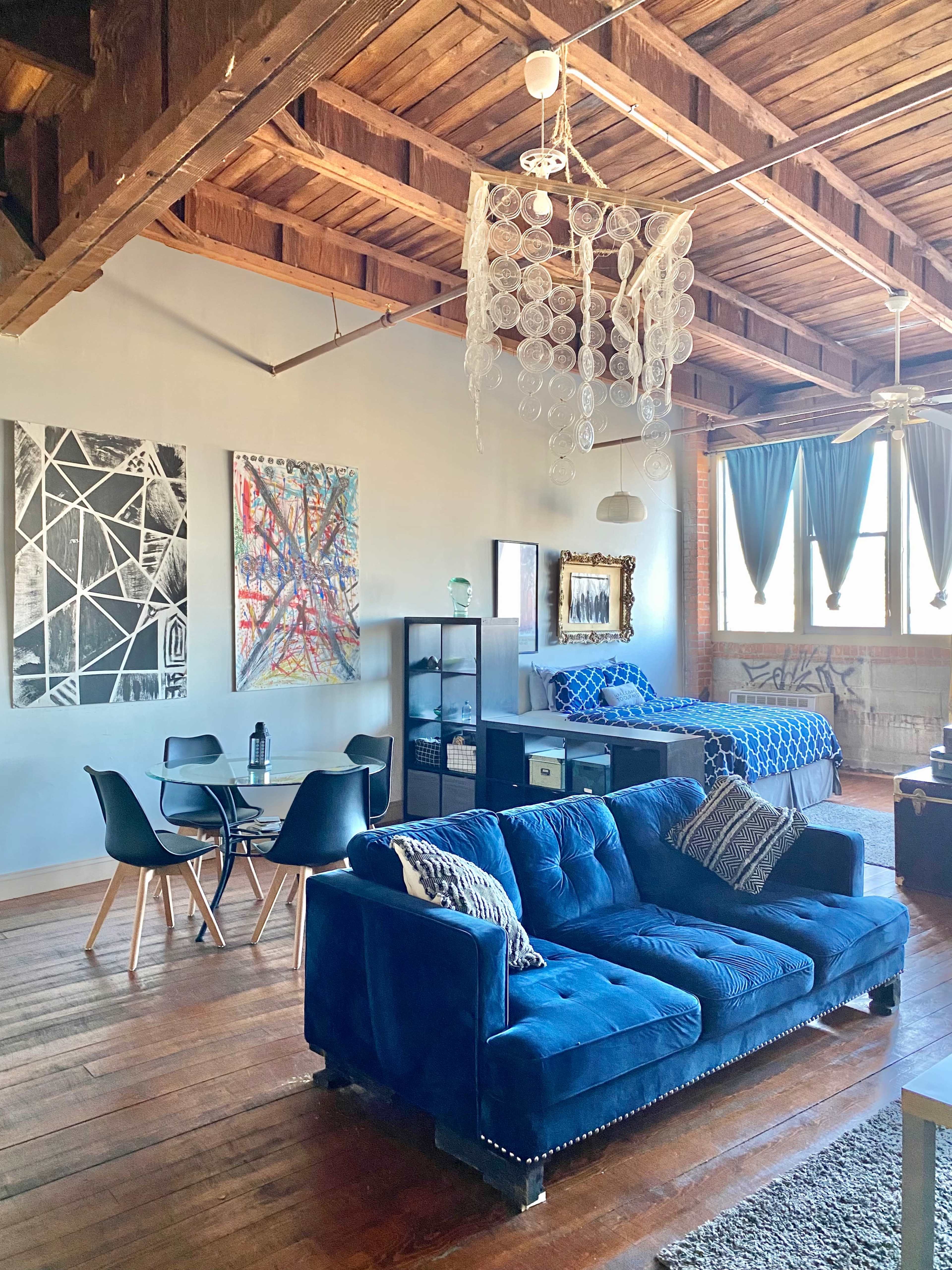 A spacious loft with wooden beams, featuring a blue couch, a circular dining table, and a bed area adorned with blue bedding.