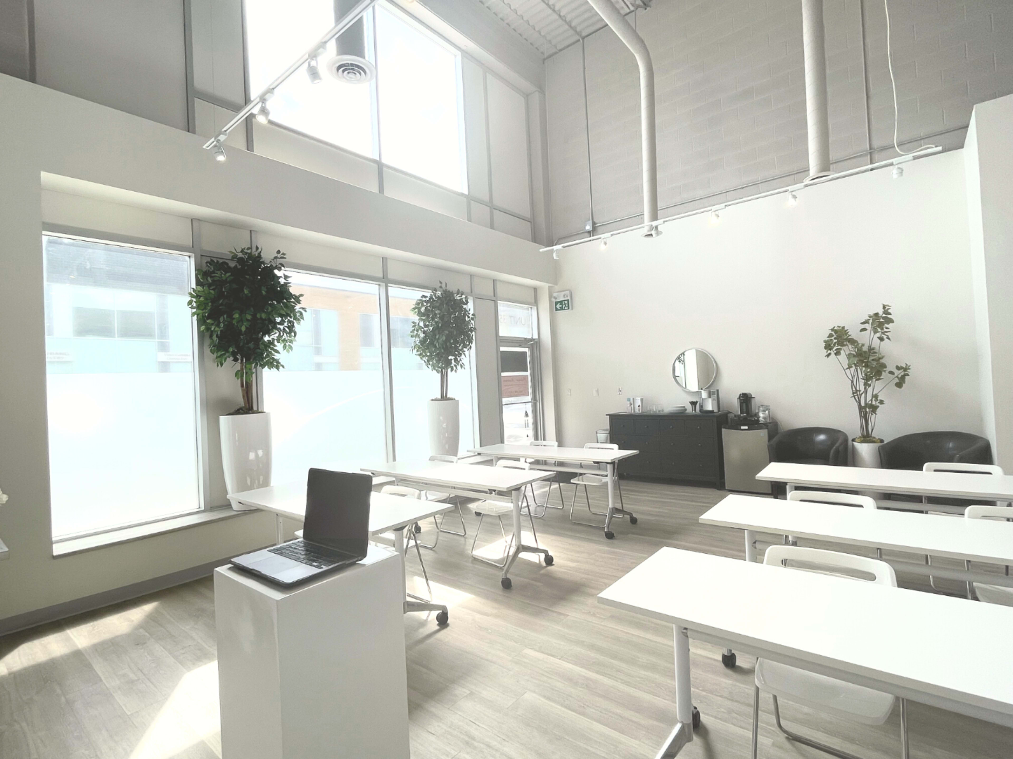 A bright, modern classroom equipped with white tables, chairs, and a large window with greenery.