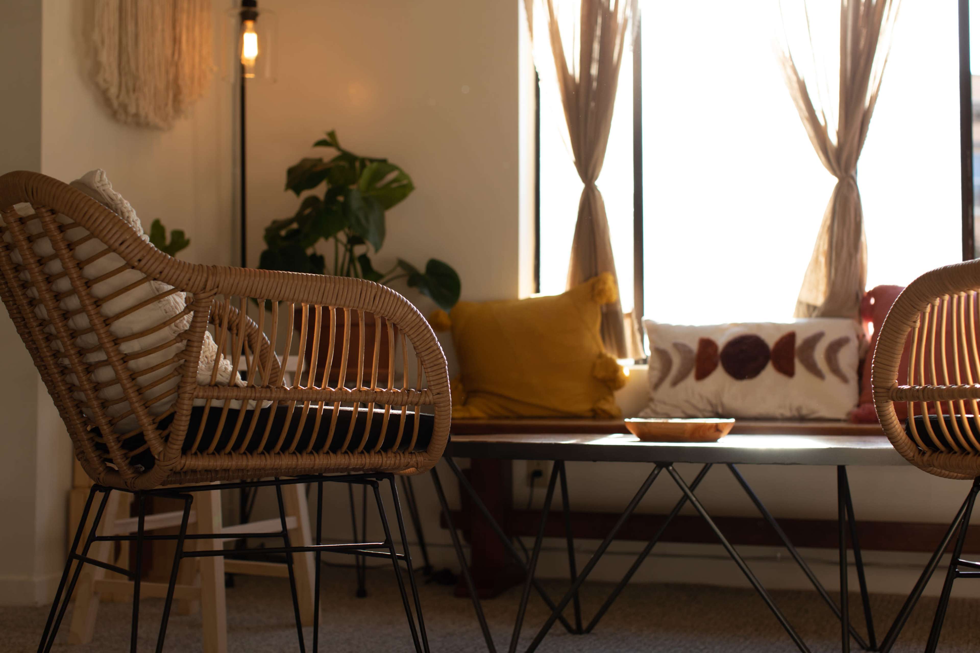 A cozy living space with wicker chairs, a wooden table, and bright natural light streaming through large windows adorned with curtains.