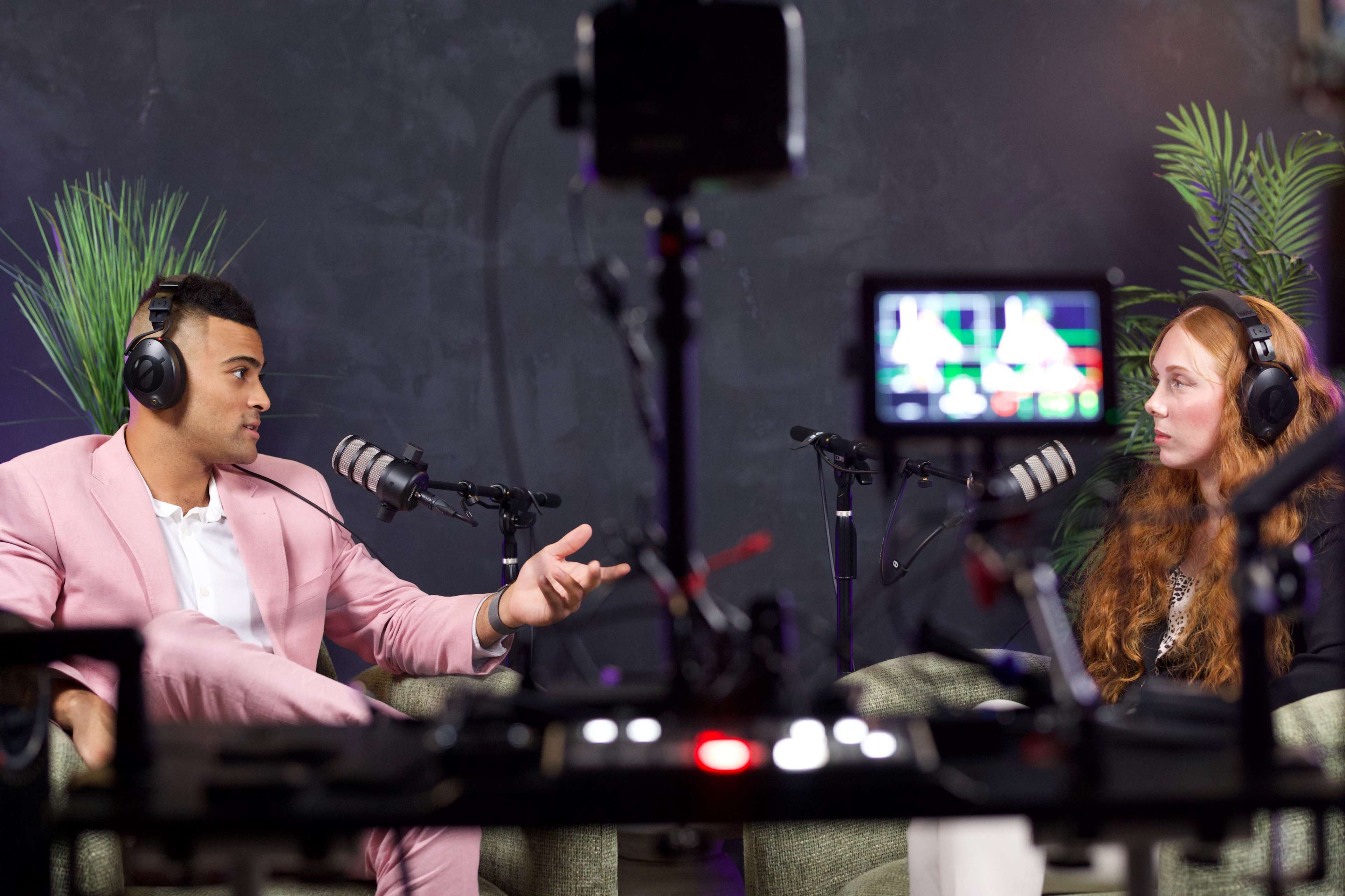 A man in a pink suit and a woman with red hair are engaged in a conversation while wearing headphones in a studio setting.