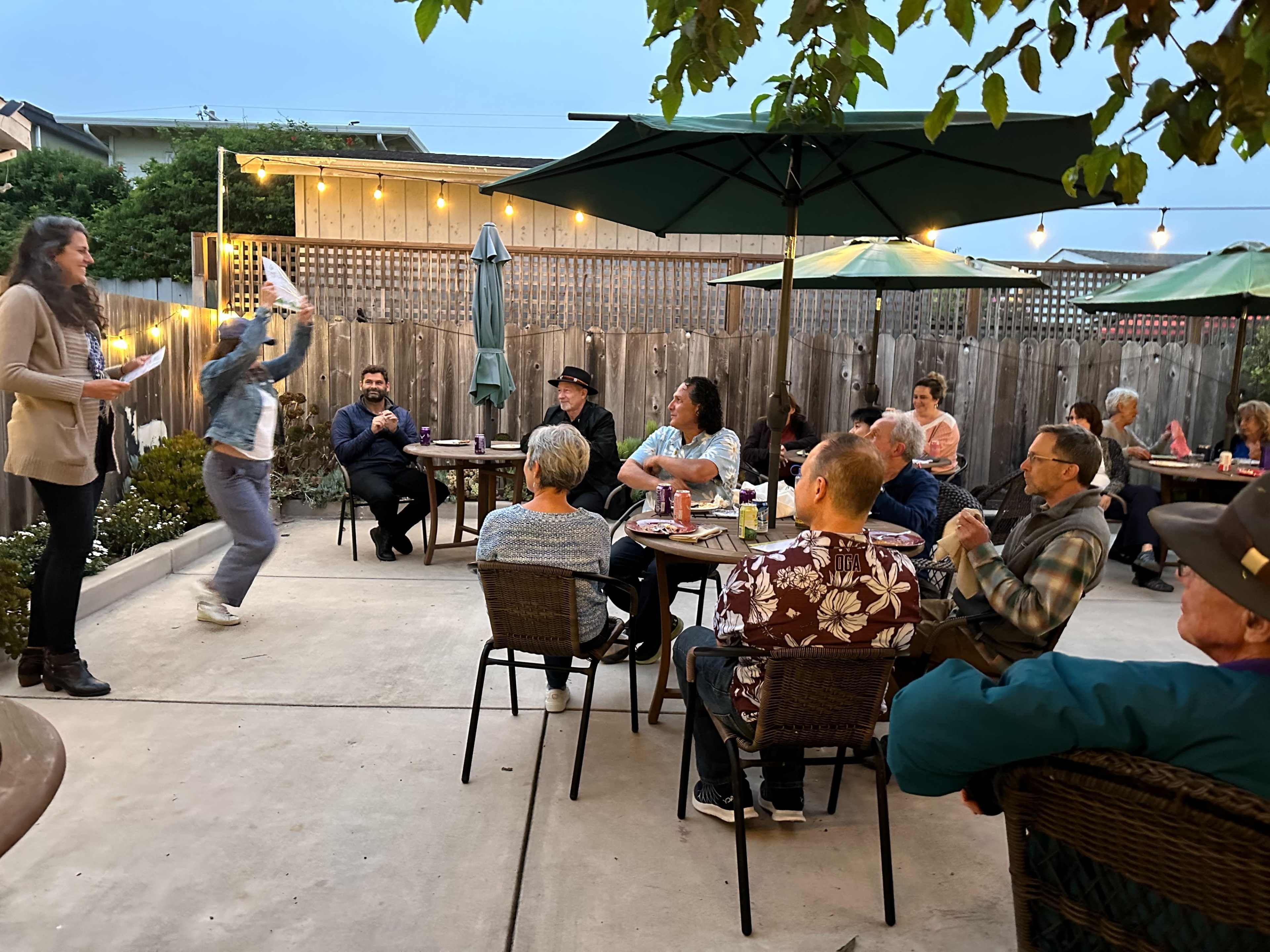 A group of people sits at tables in an outdoor patio setting, while one person performs in front of them under evening lights.