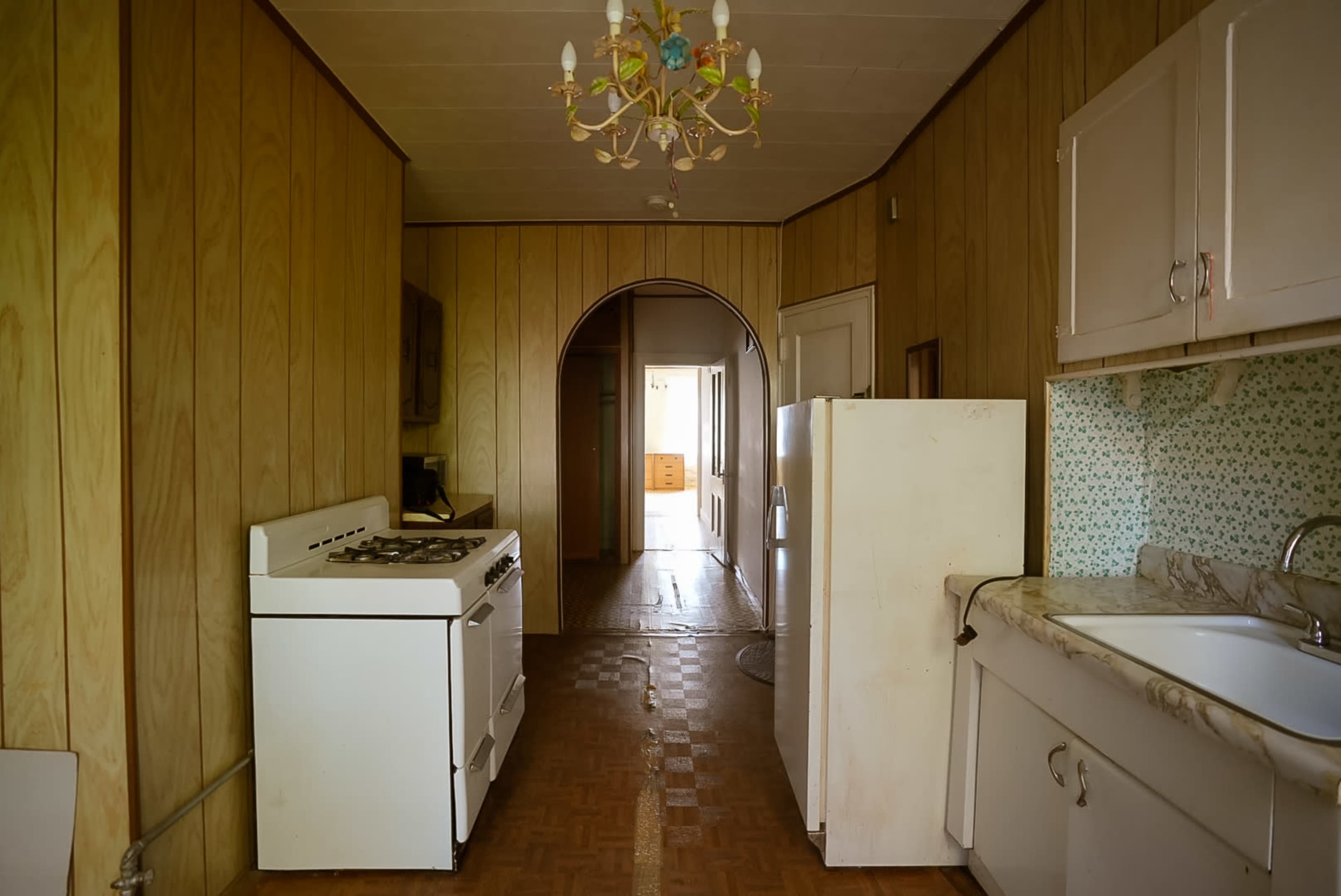 The image shows a narrow kitchen with wooden paneling, featuring a white stove, refrigerator, and a sink, leading to a hallway.