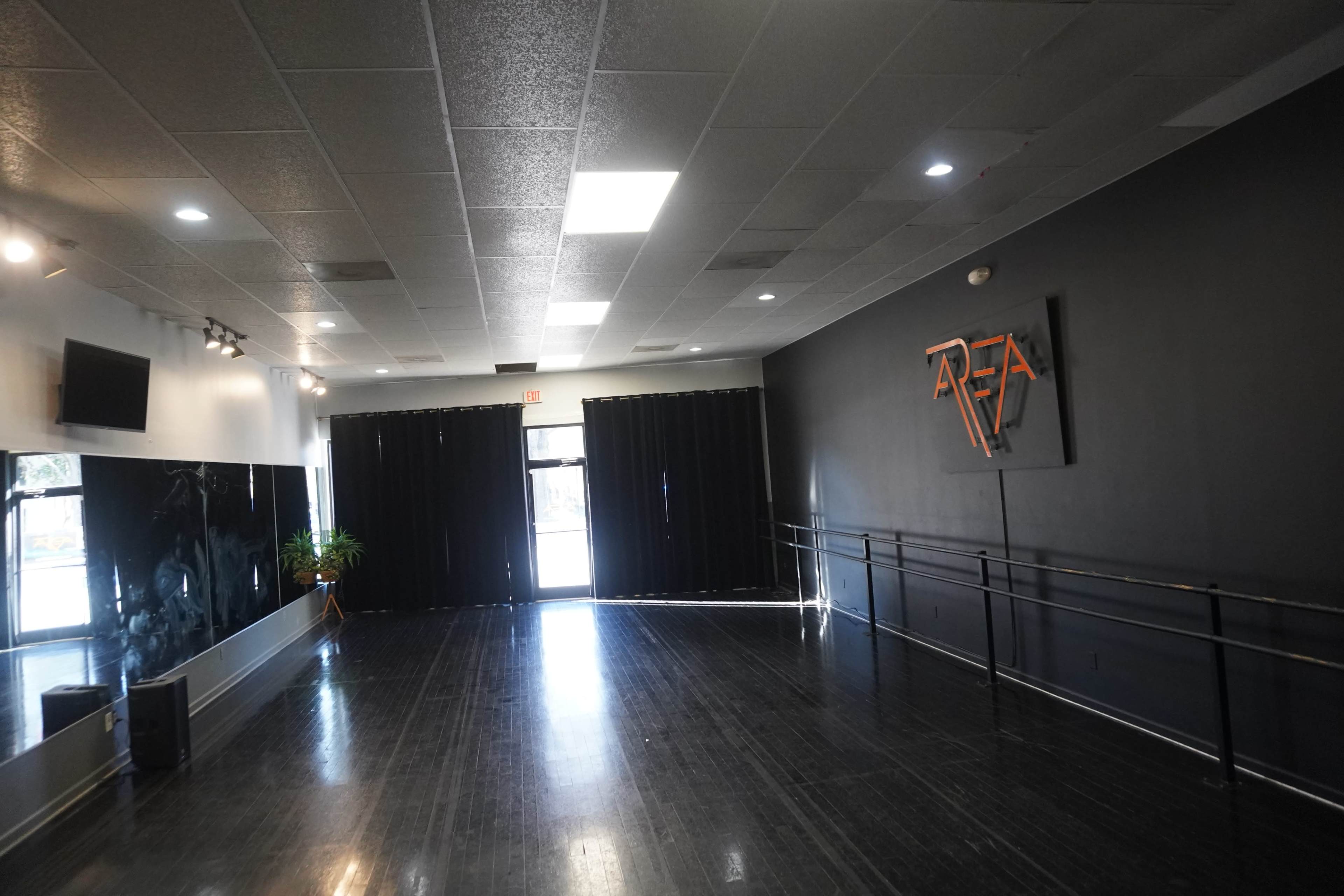 An empty dance studio with black walls, wooden flooring, a wall-mounted mirror, and a logo on the wall.