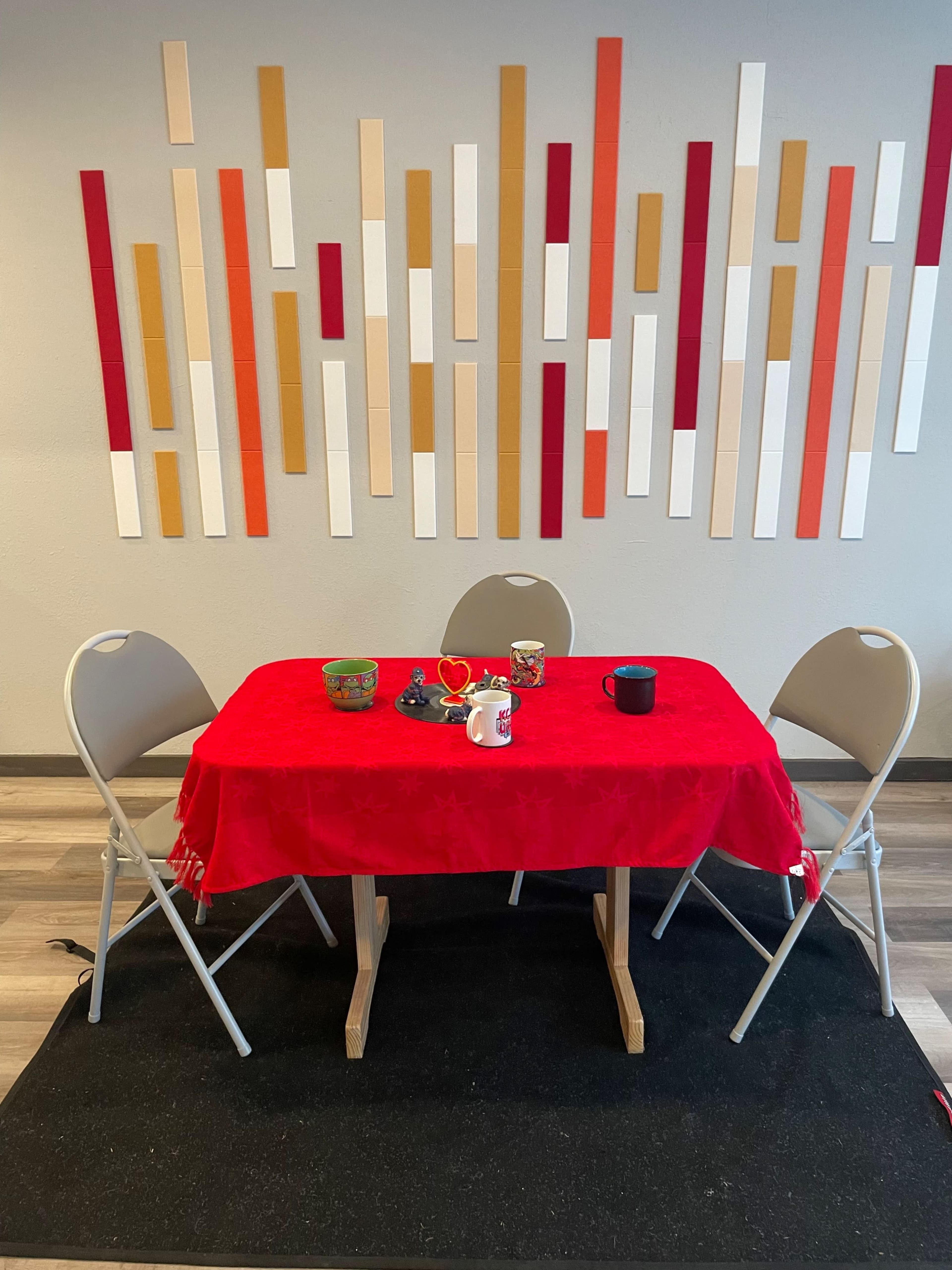 A small table with a red tablecloth and three folding chairs is set against a wall decorated with vertical colored strips.