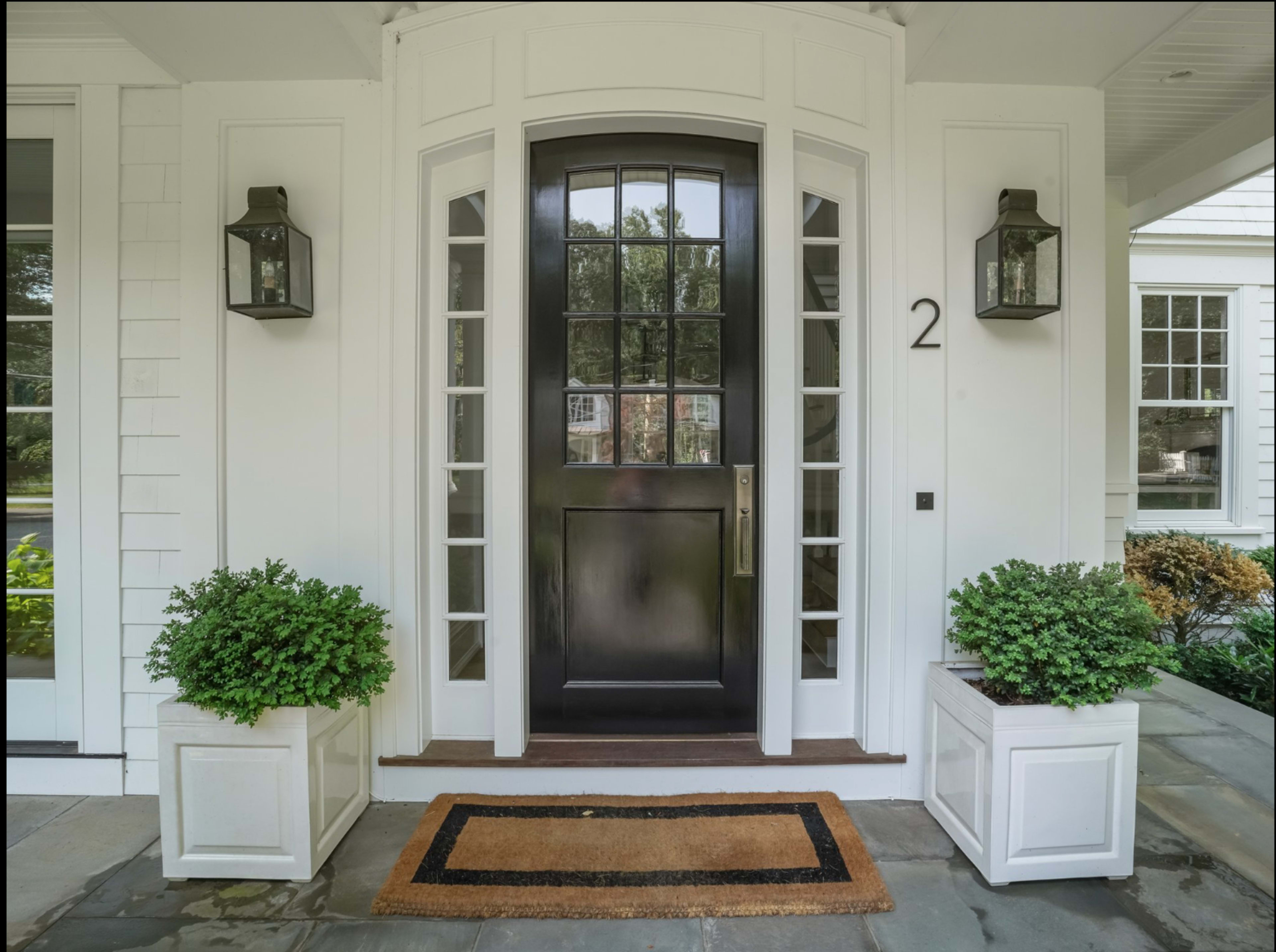 The image shows a front entrance featuring a black door with glass panels, flanked by two lanterns and greenery in white planters, accompanied by a welcome mat.