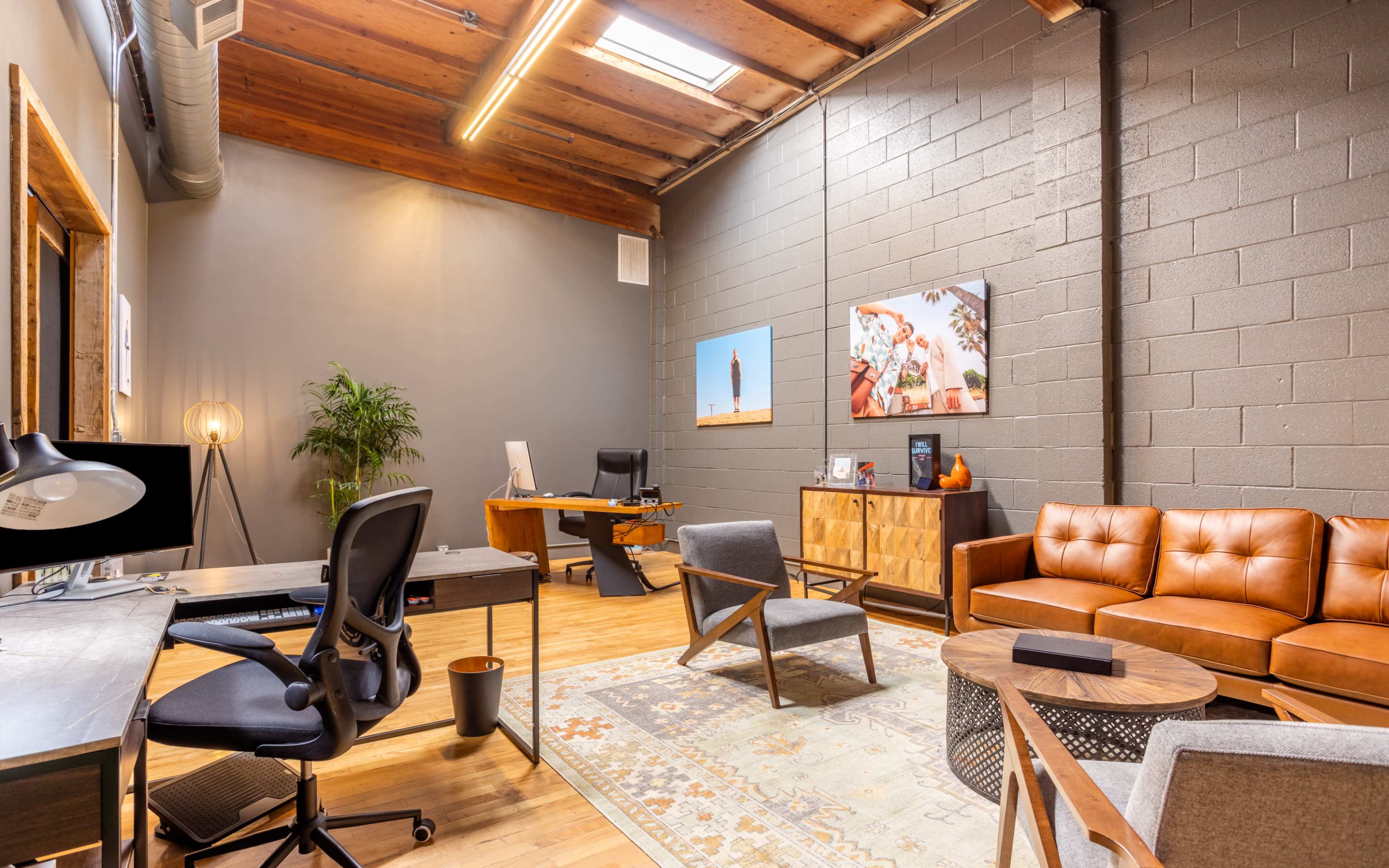 A modern office space featuring a wooden desk and chairs, a leather sofa, a large window with natural light, and decorative wall art.