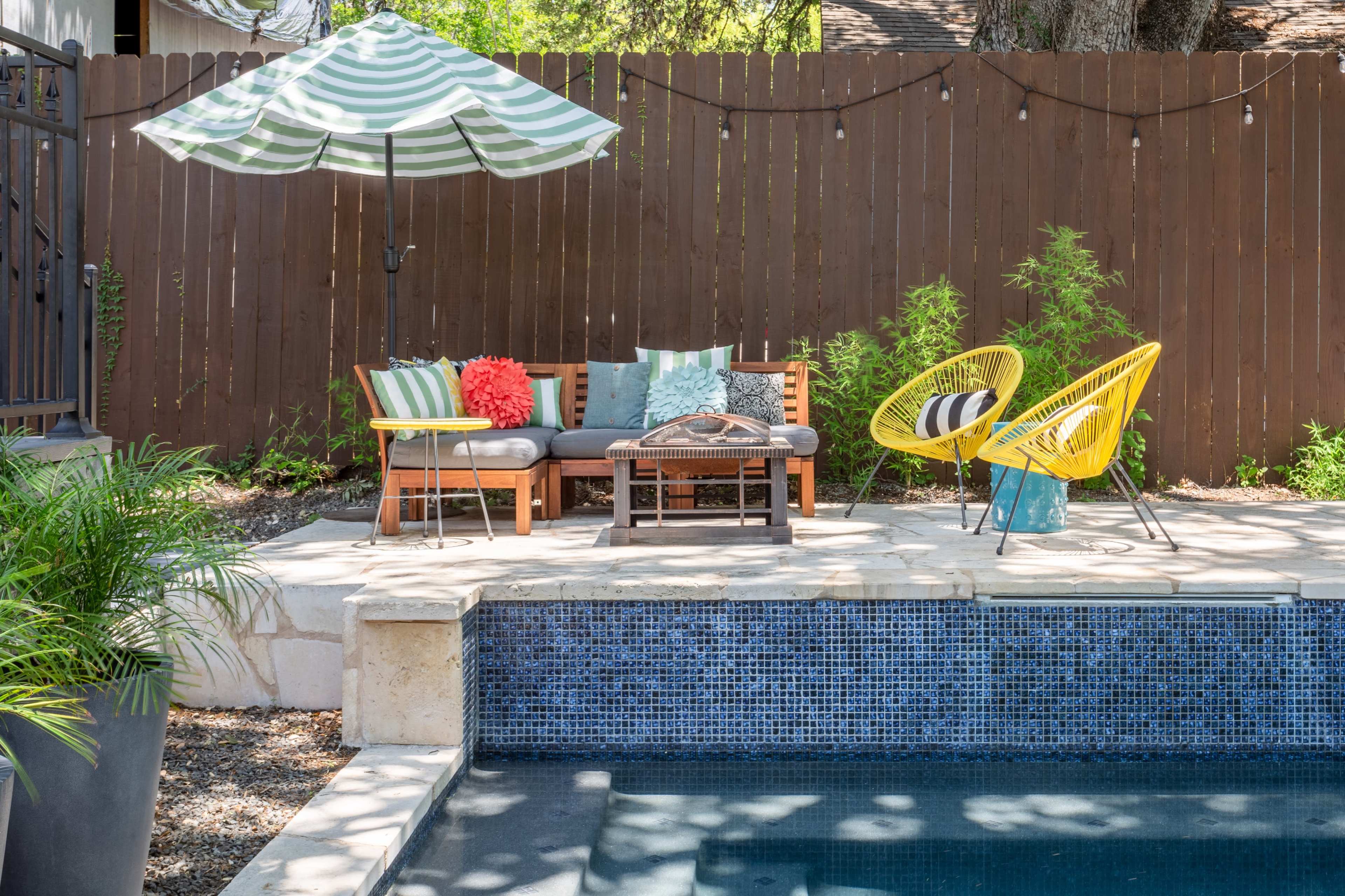 The scene features a poolside area with a wooden lounge set, a striped umbrella, and colorful chairs beside a blue-tiled pool.