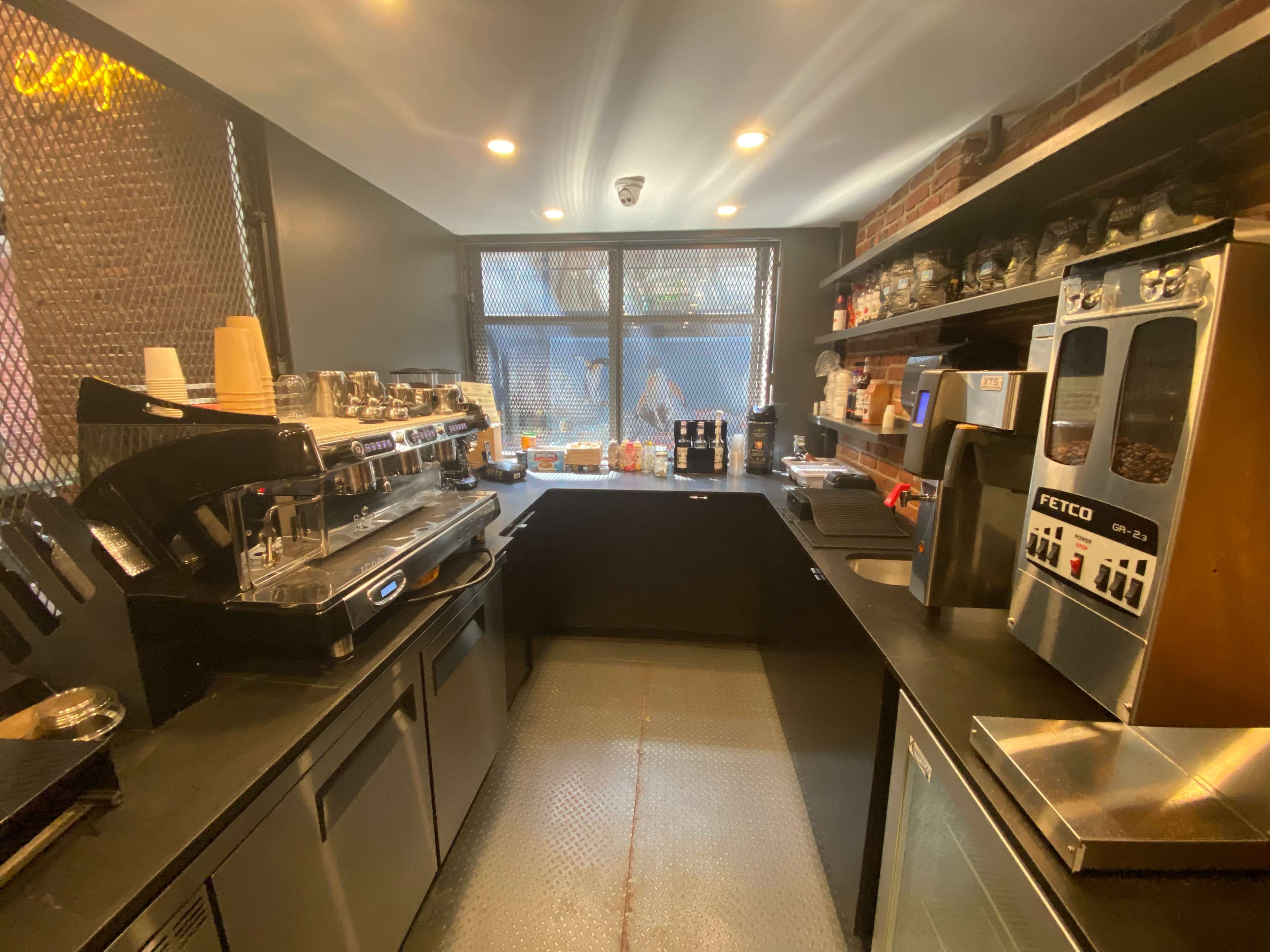 A compact coffee shop kitchen with equipment including an espresso machine, coffee grinder, and shelves stocked with jars and utensils.