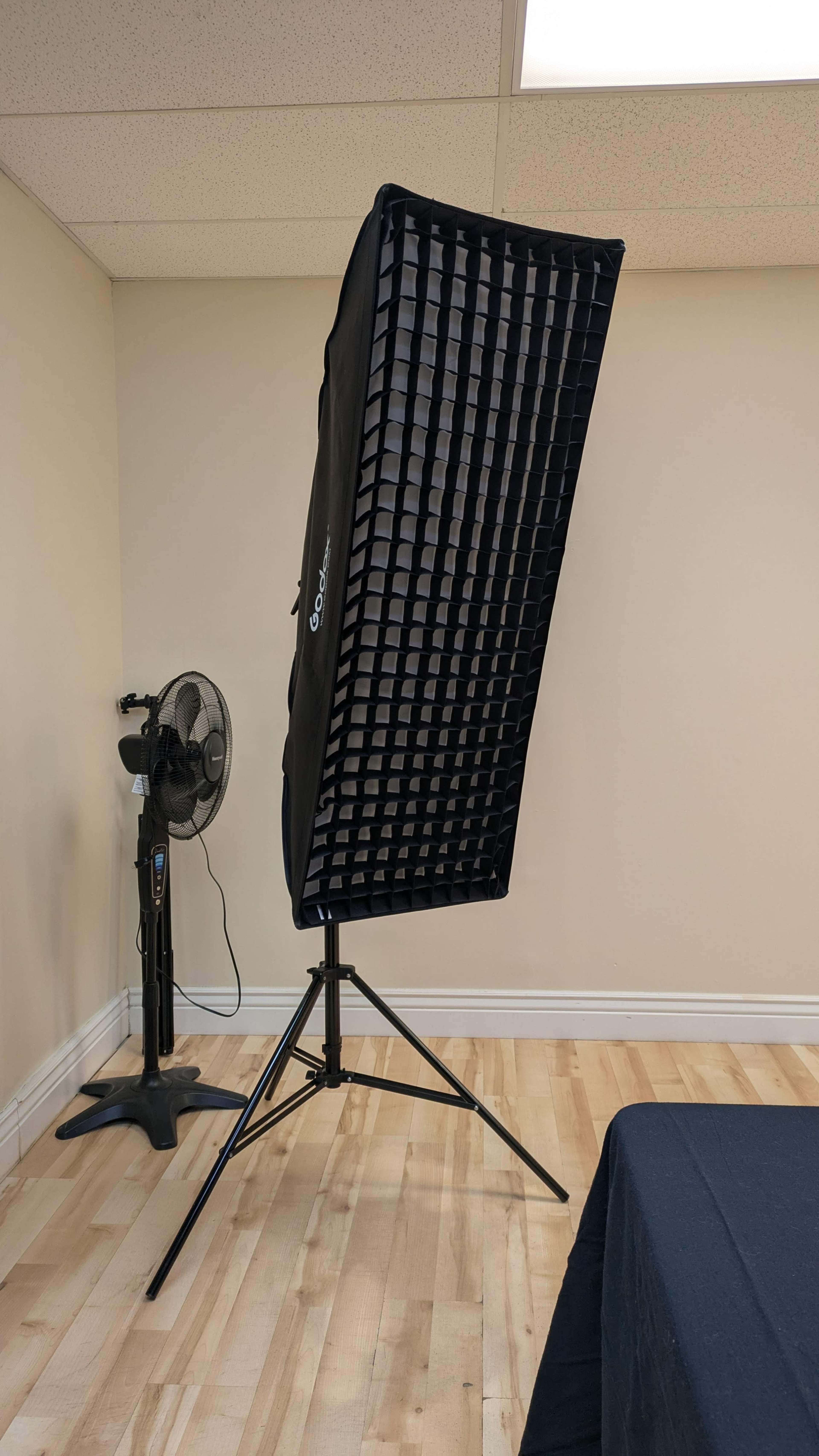 A large photography softbox is positioned on a stand next to a fan in a room with wooden flooring and light-colored walls.