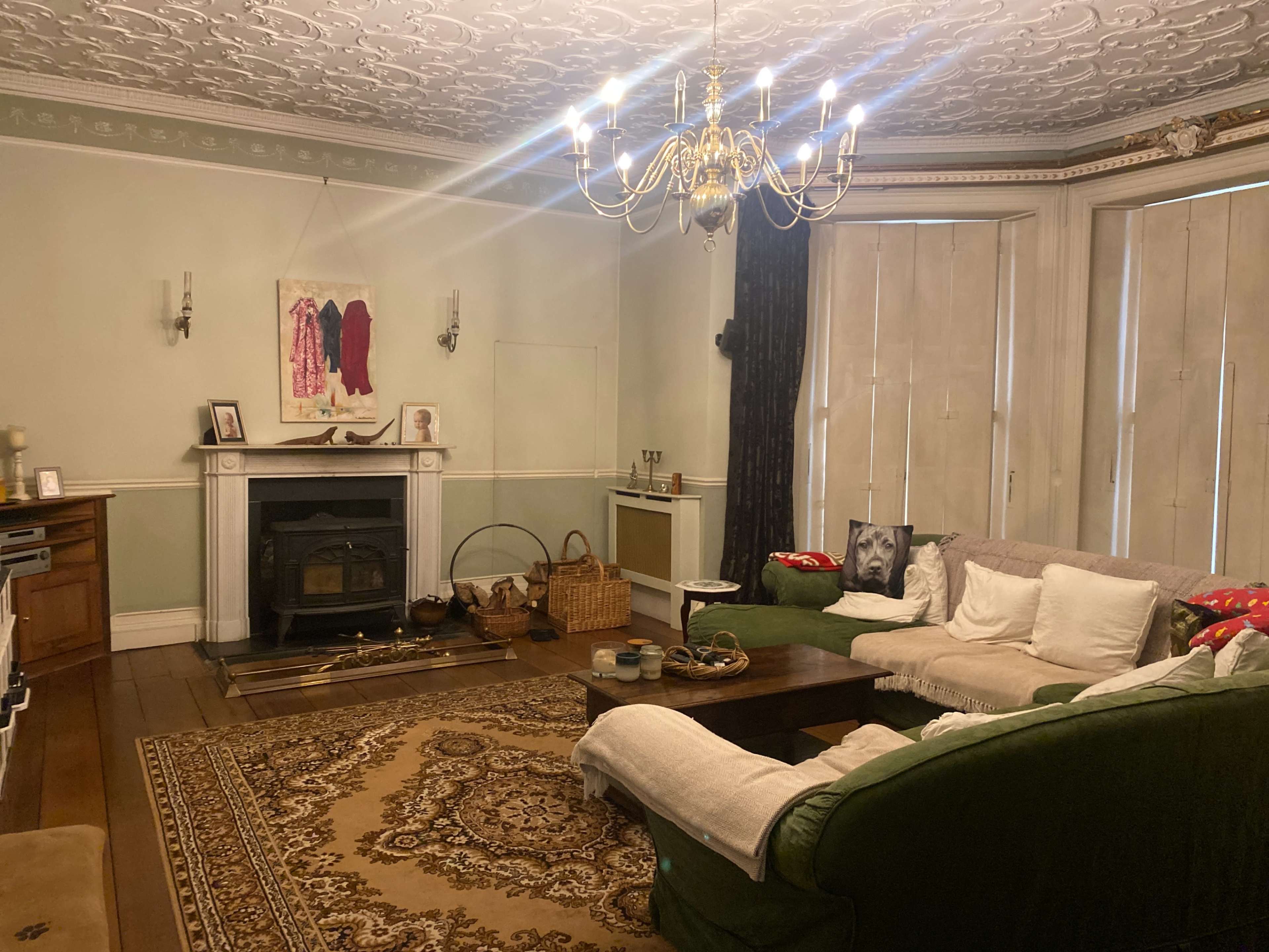 A cozy living room with a green sofa, a wooden coffee table, and a decorative chandelier, featuring a patterned rug and large, curtain-covered windows.