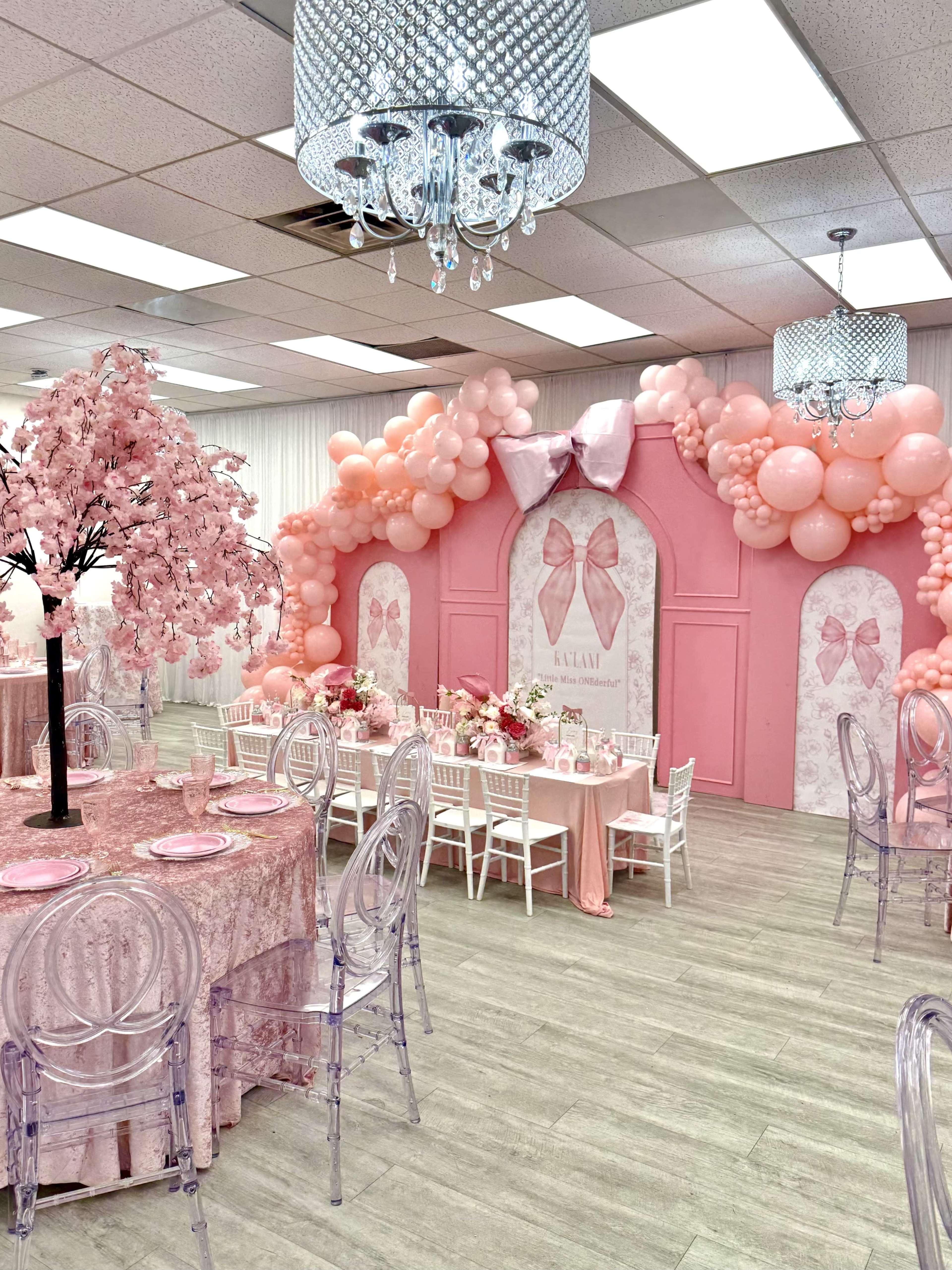 The image shows a decorated event space featuring pink-themed decorations, including a balloon arch, a pink backdrop with bows, and tables set with floral arrangements.