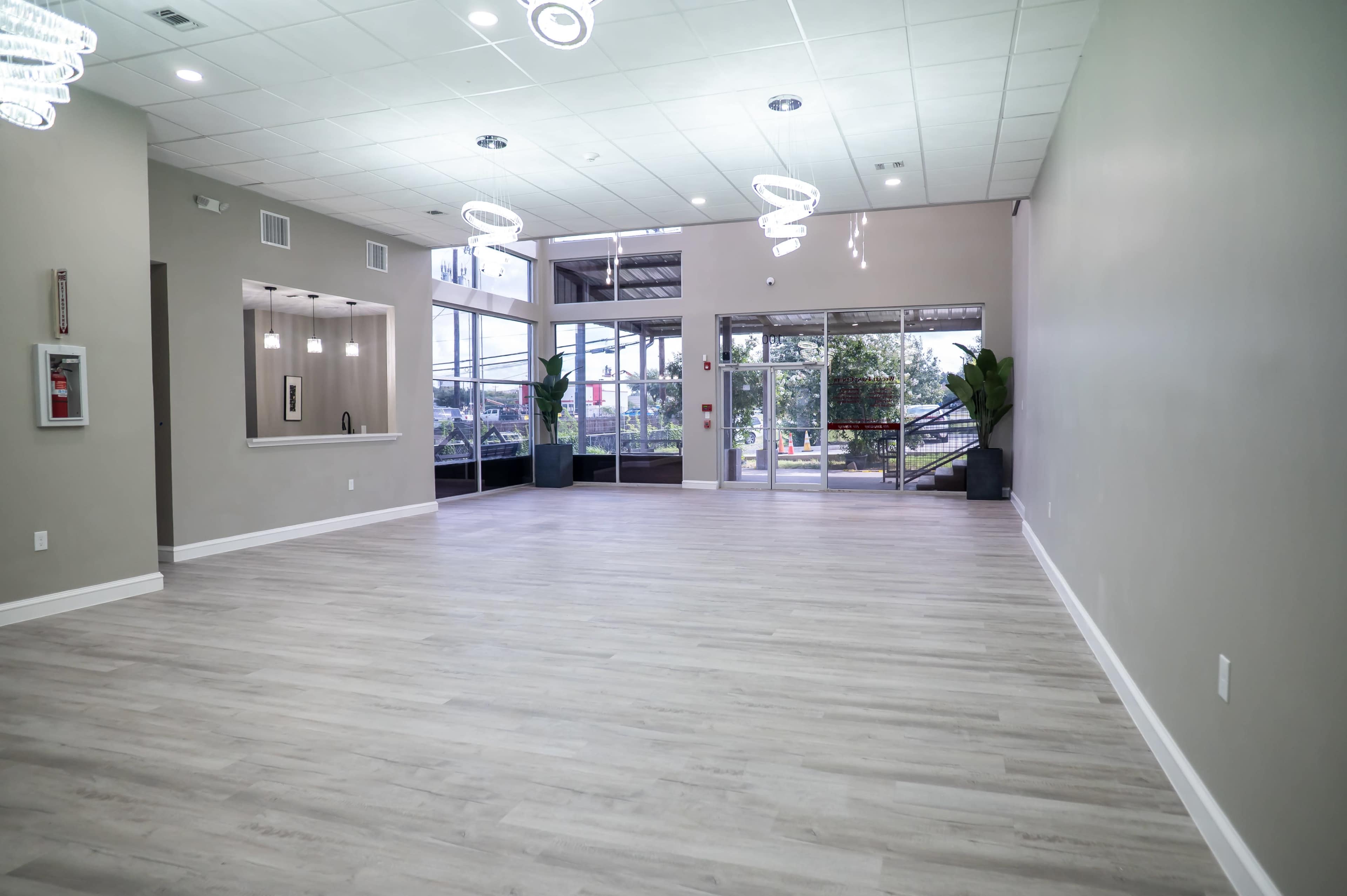 The image shows an empty, modern interior space with wooden flooring, large windows, and minimal decorative elements.