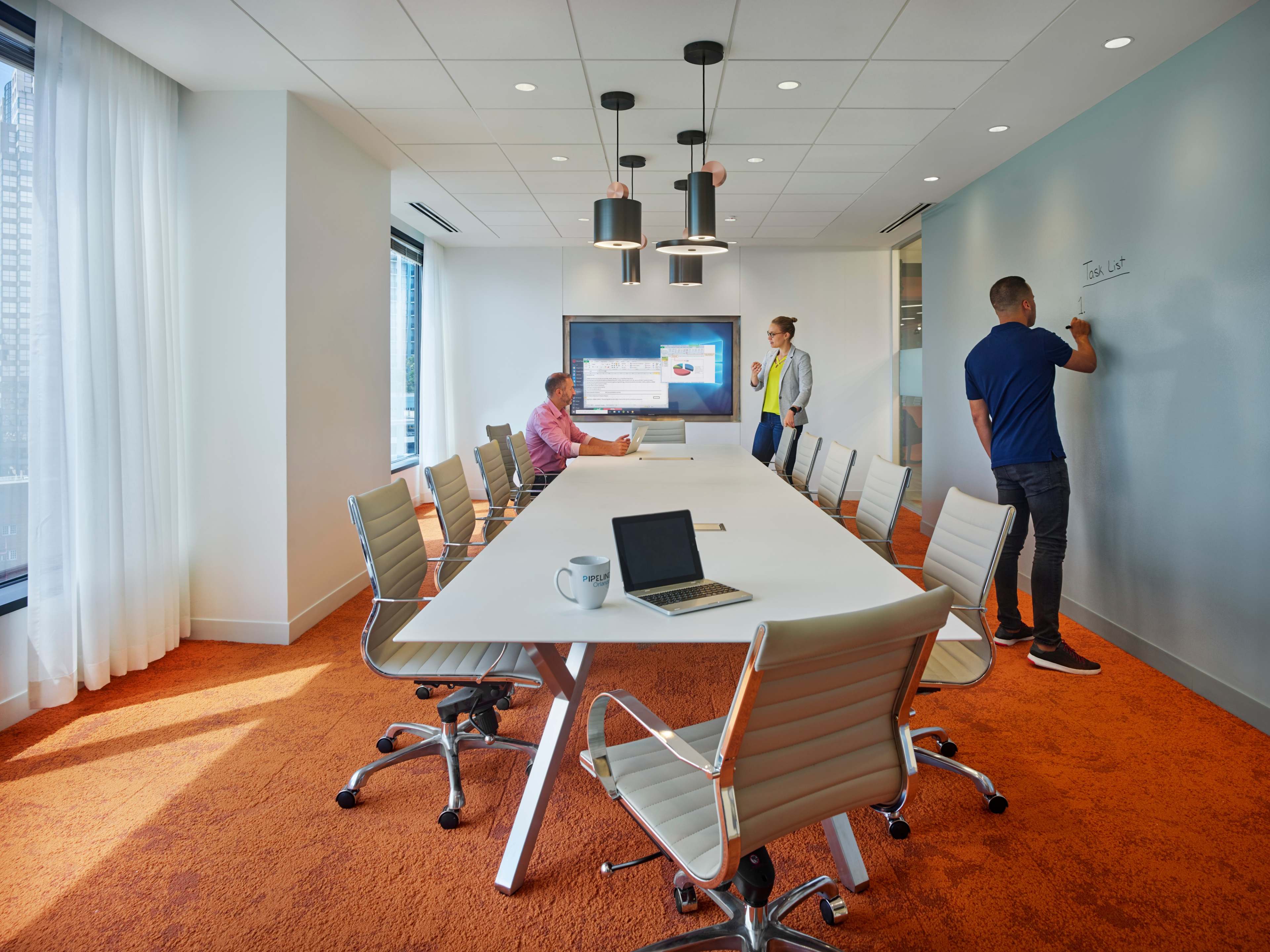 A modern conference room features a long table surrounded by ergonomic chairs, with a large screen displaying a presentation and two individuals writing on a wall.