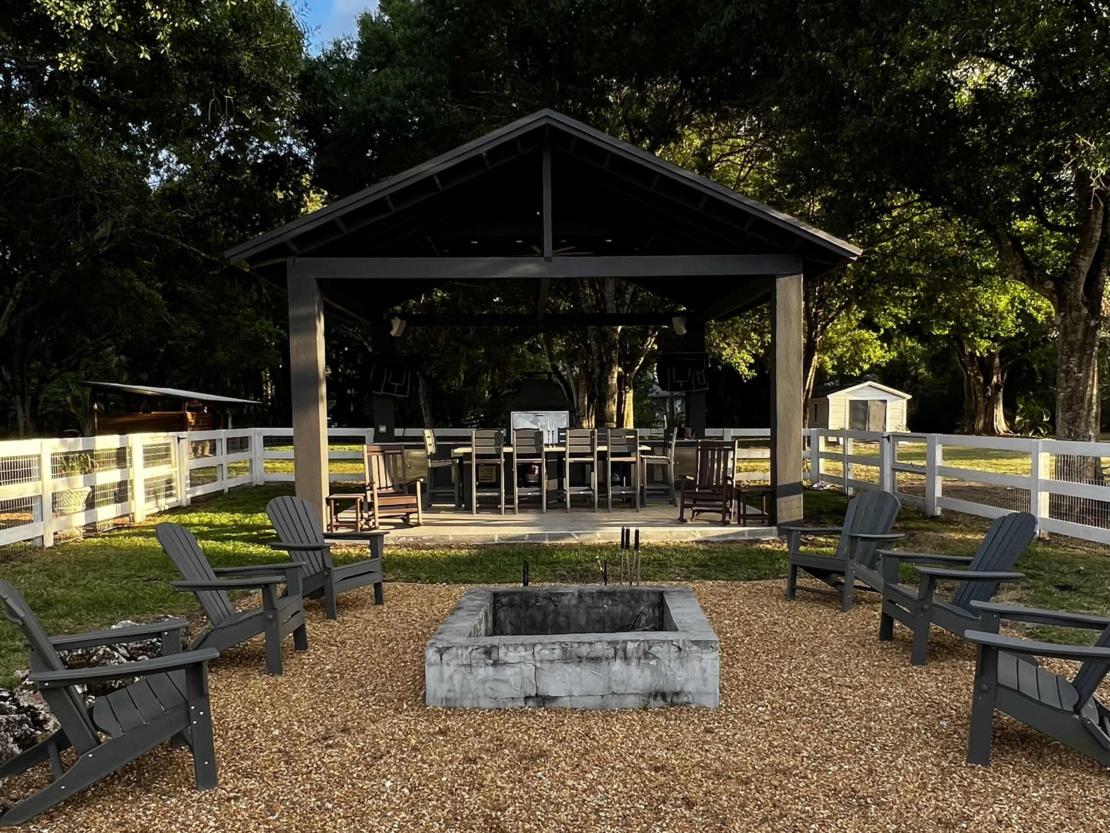A stone fire pit surrounded by gray Adirondack chairs sits in front of a covered seating area with a large table and chairs, all framed by trees and a white fence.