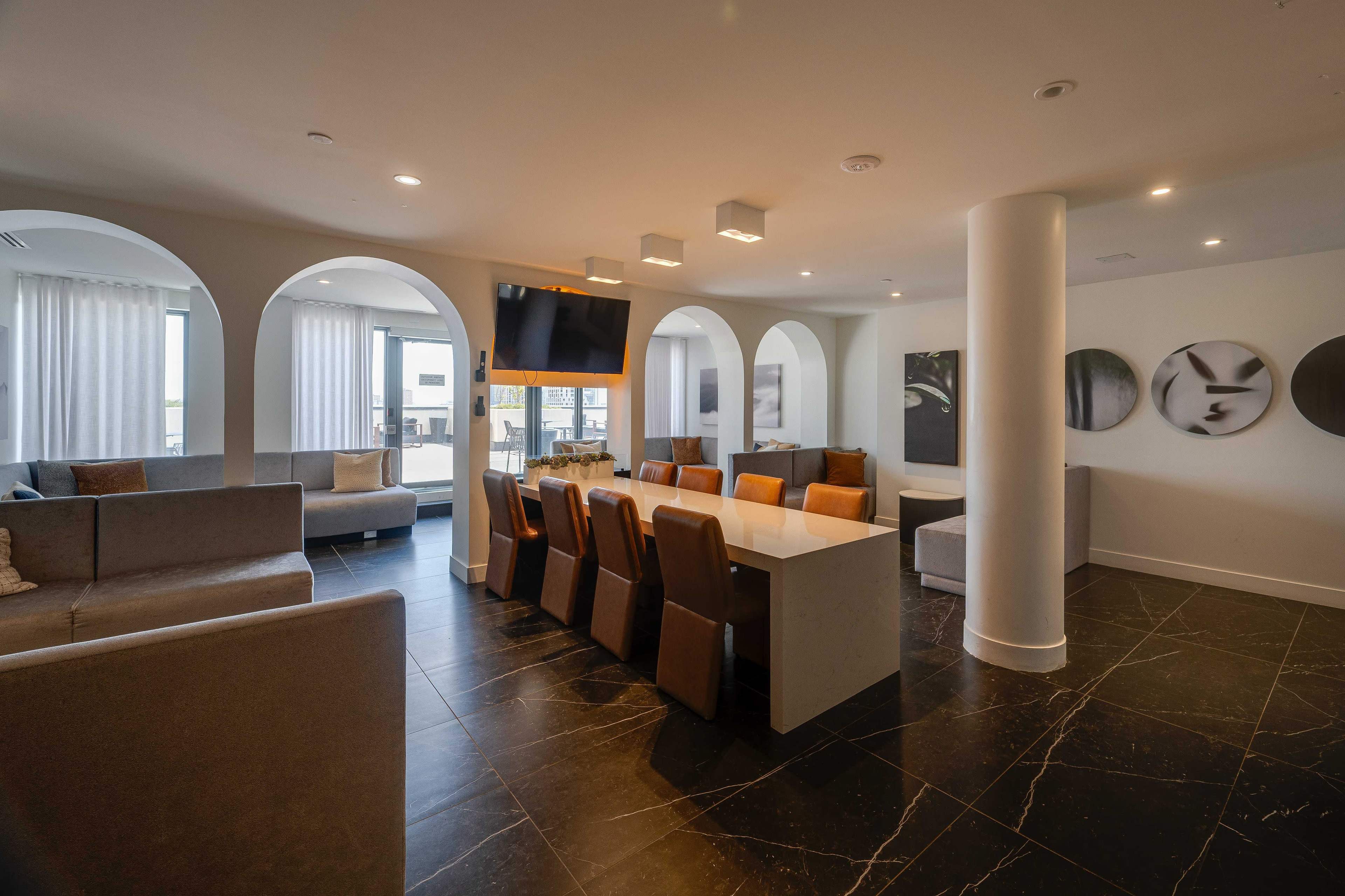 The image shows a modern interior space with a dining area featuring a large white table and brown chairs, surrounded by soft seating areas and large windows.