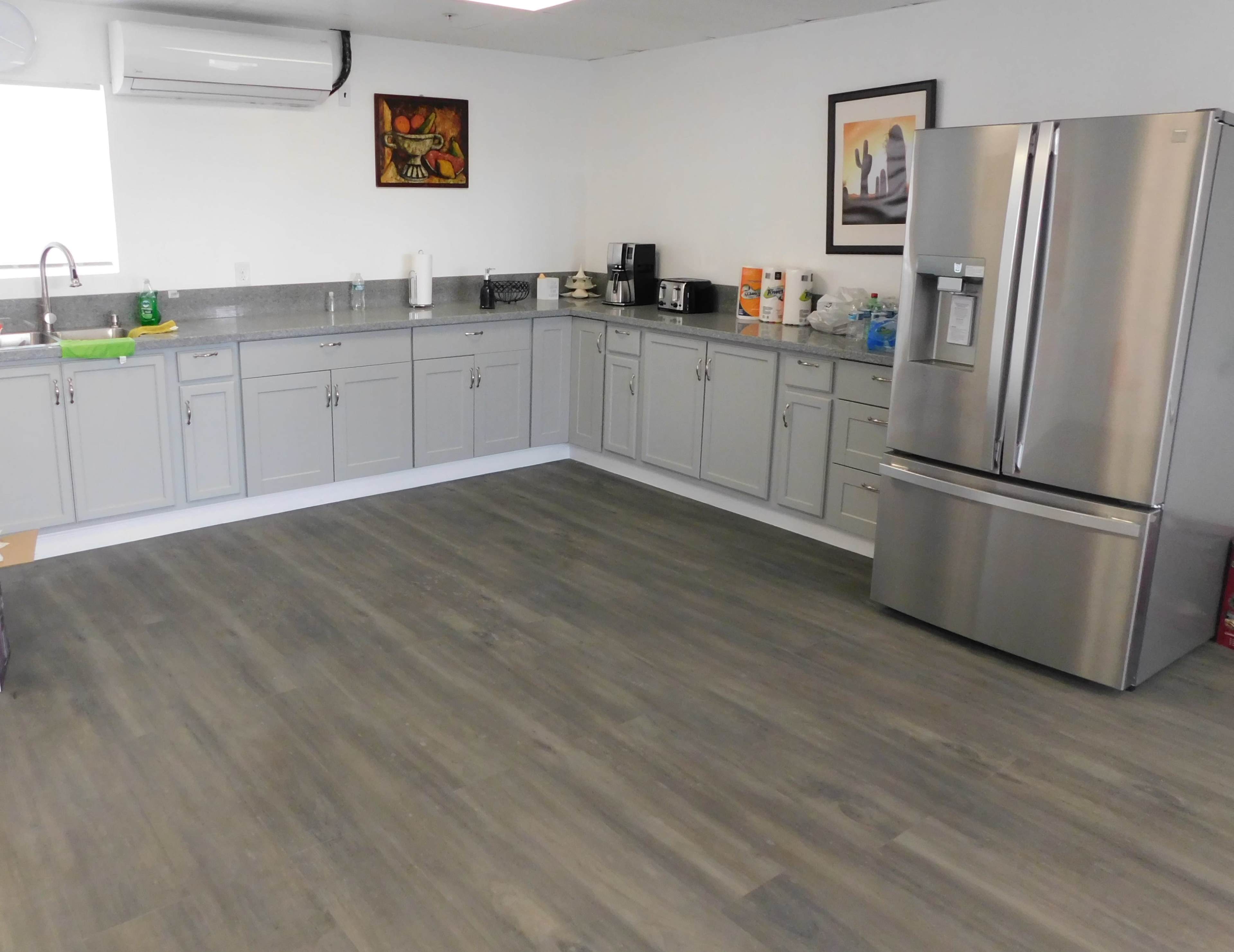 The image shows a spacious kitchen with gray cabinets, a stainless steel refrigerator, and various kitchen appliances arranged along the walls.