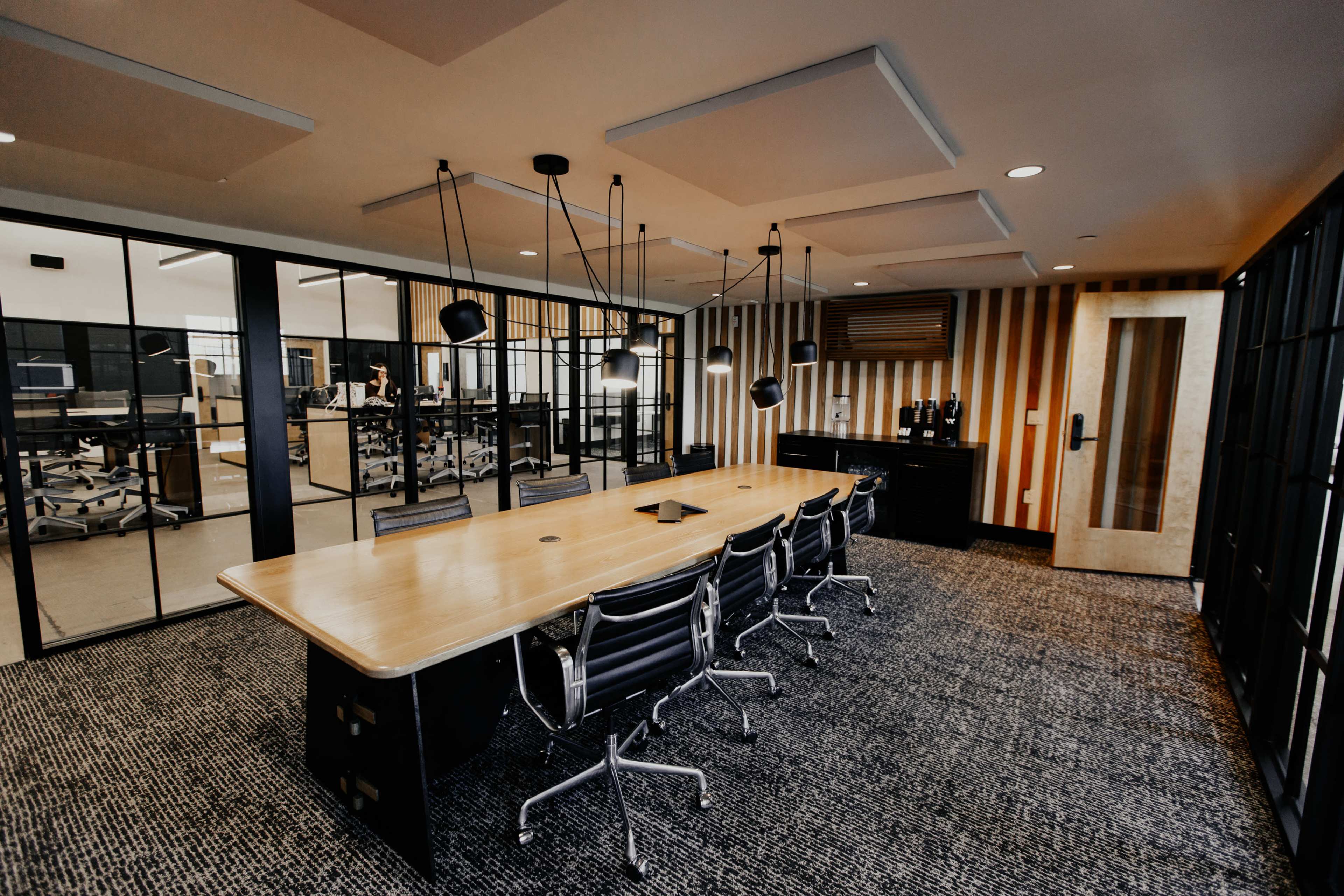 A modern conference room featuring a long wooden table surrounded by black chairs, with glass walls and pendant lighting overhead.