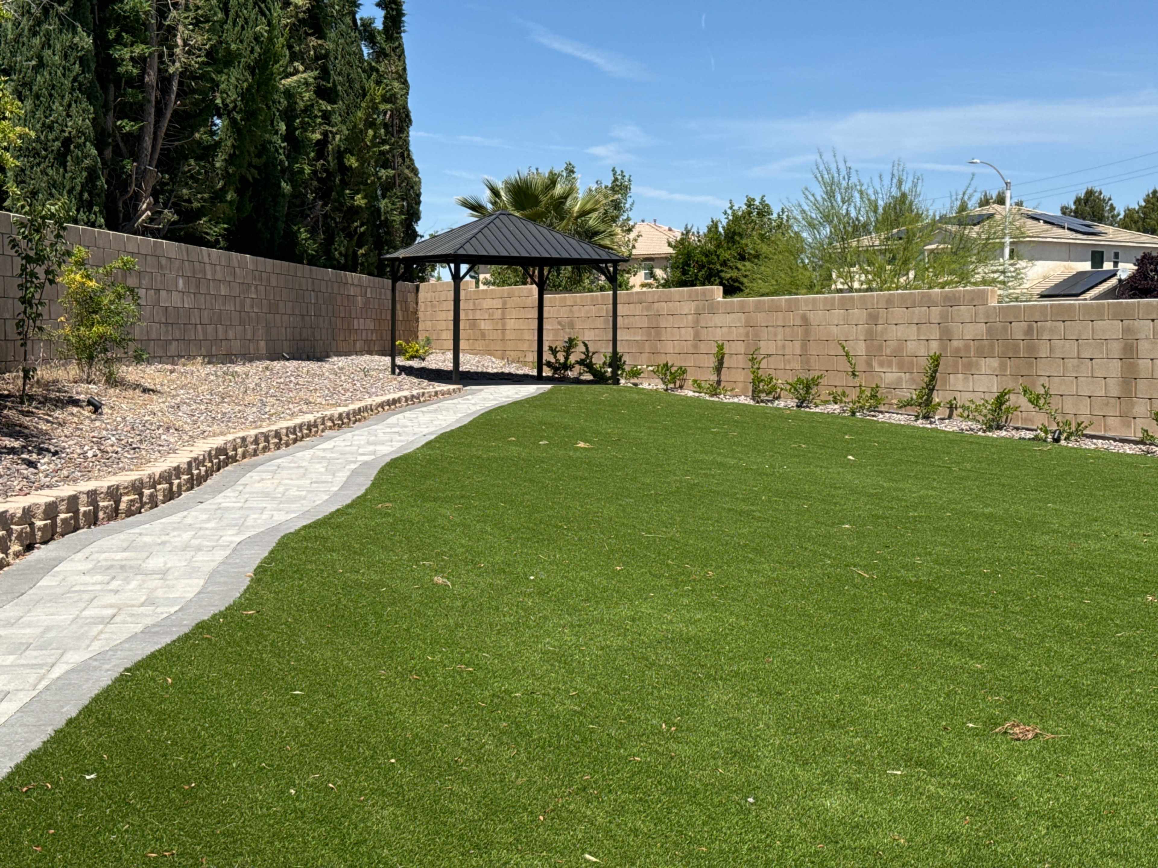 The image shows a landscaped backyard with artificial turf, a stone pathway, and a gazebo in the background surrounded by walls.