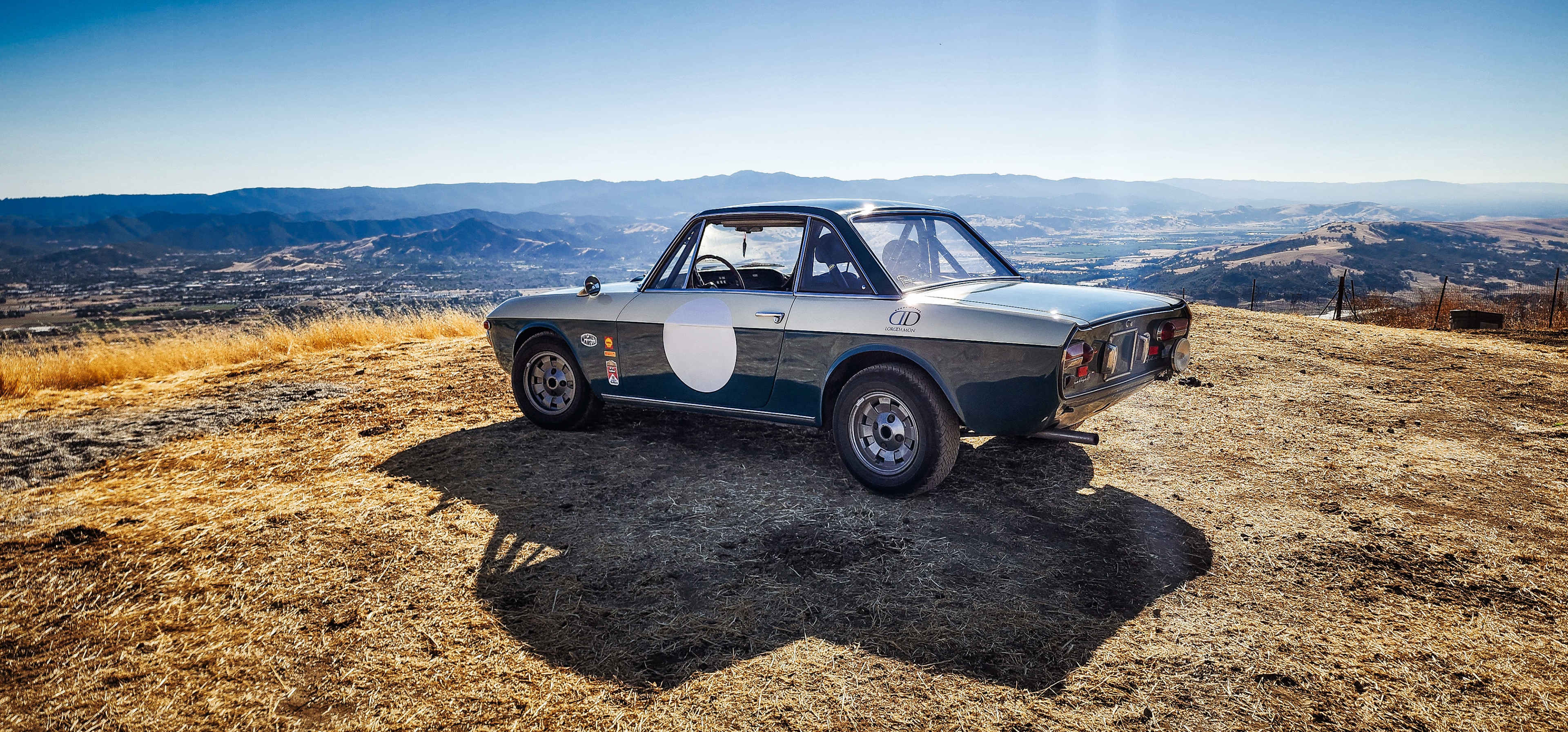 A vintage car with a circular white emblem is parked on a hillside overlooking a valley and distant mountains under a clear blue sky.