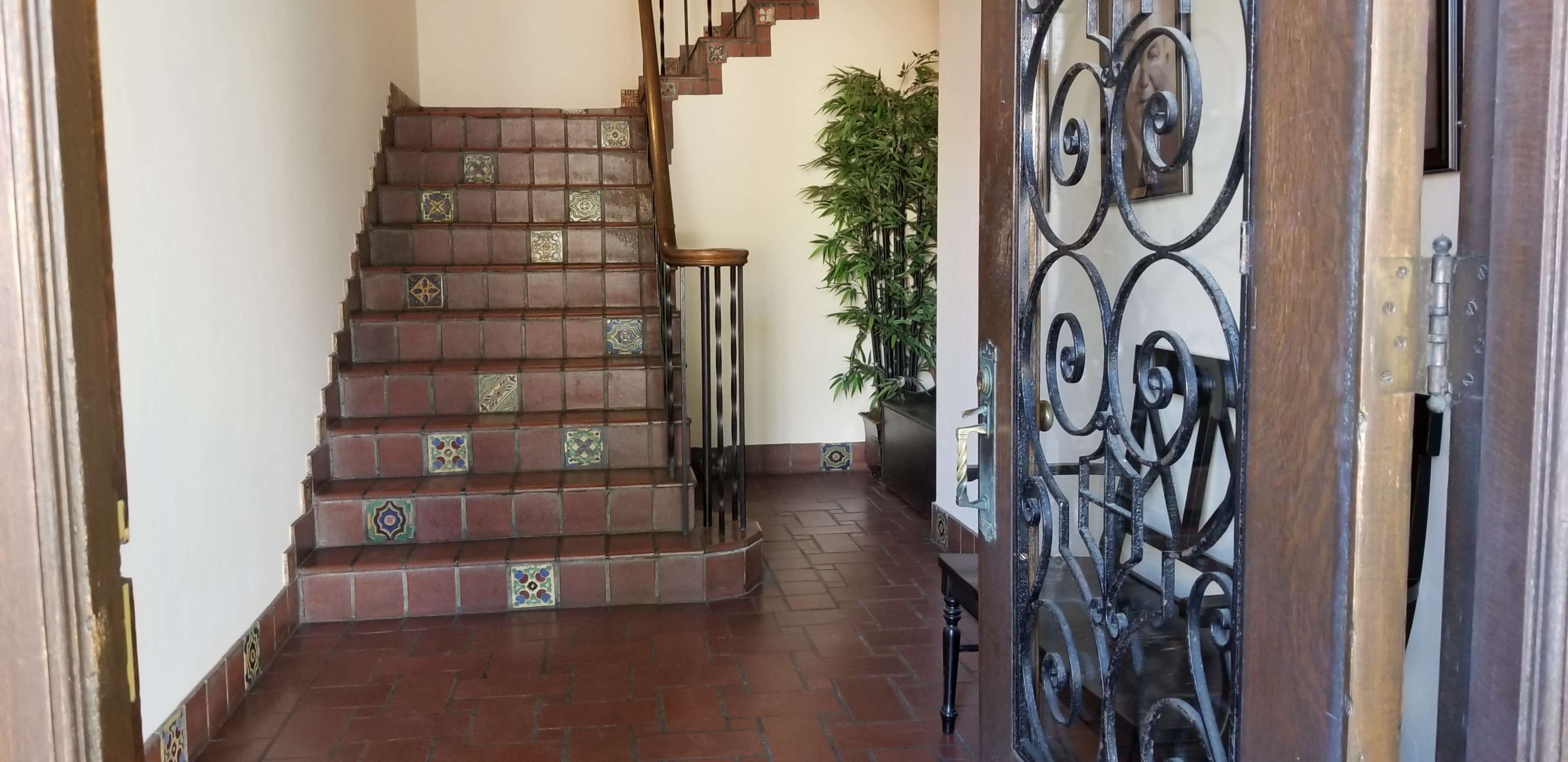 A wooden door with an ornate iron design opens to a tiled entryway featuring a staircase with decorative tiles and a potted plant.