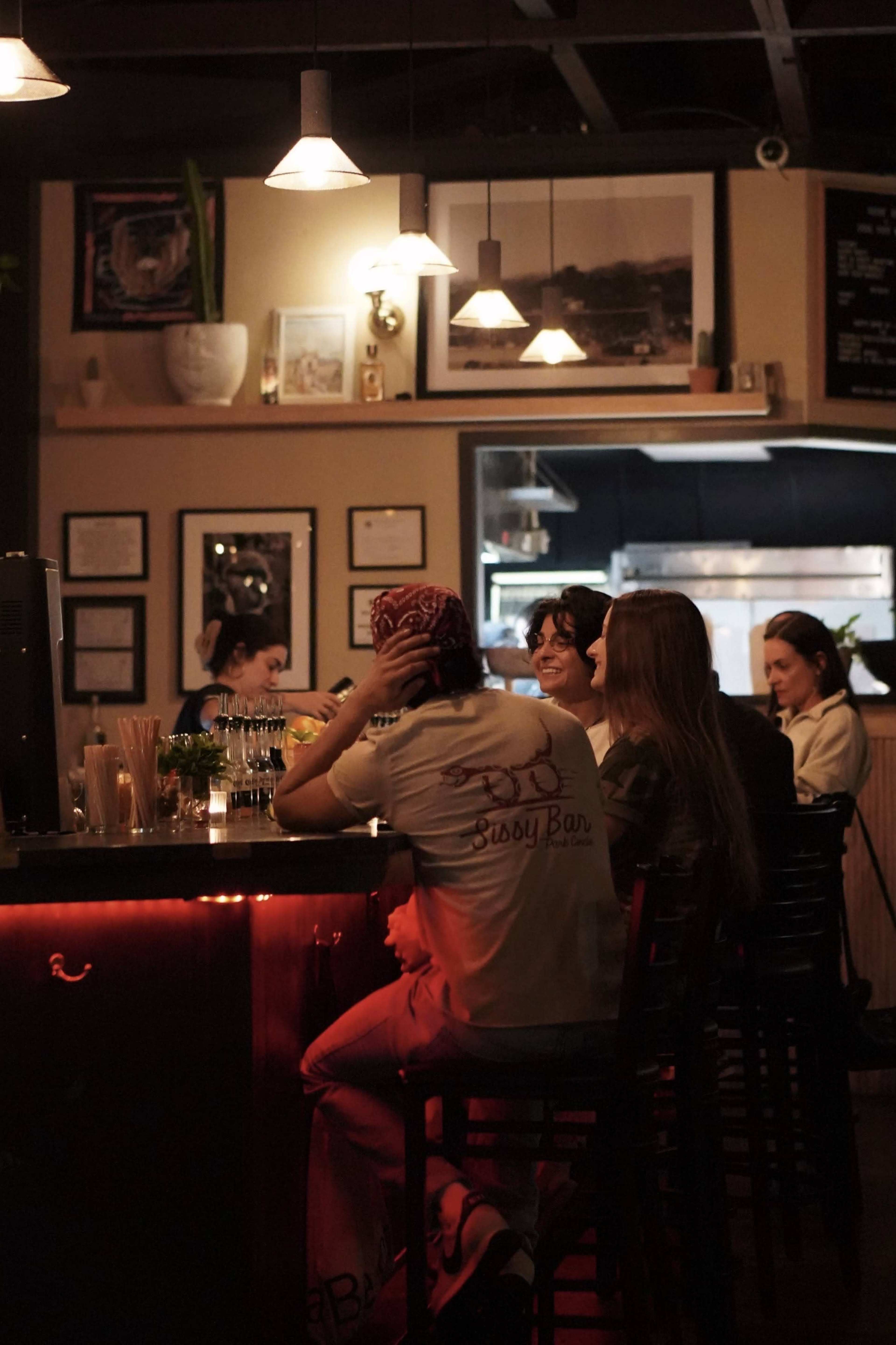 A group of patrons sits at a bar, with some looking toward the bartender while others engage in conversation.