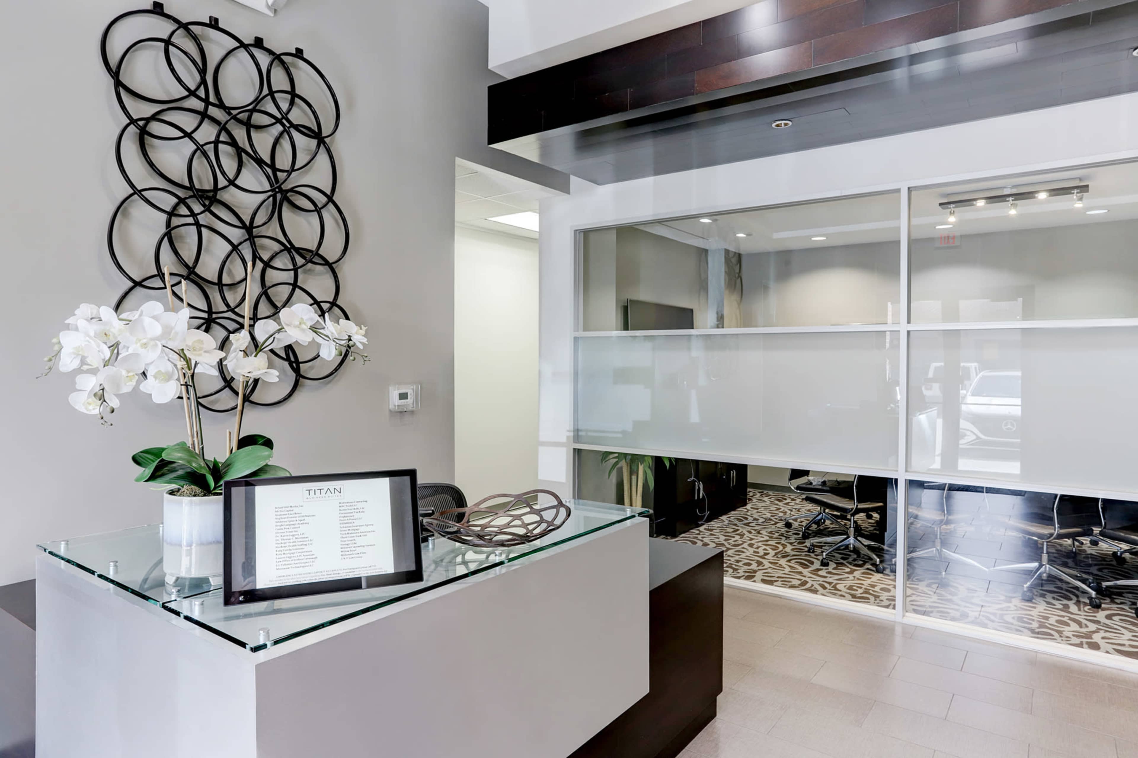 The image shows a modern office reception area with a glass counter, a plant, and a decorative wall piece.