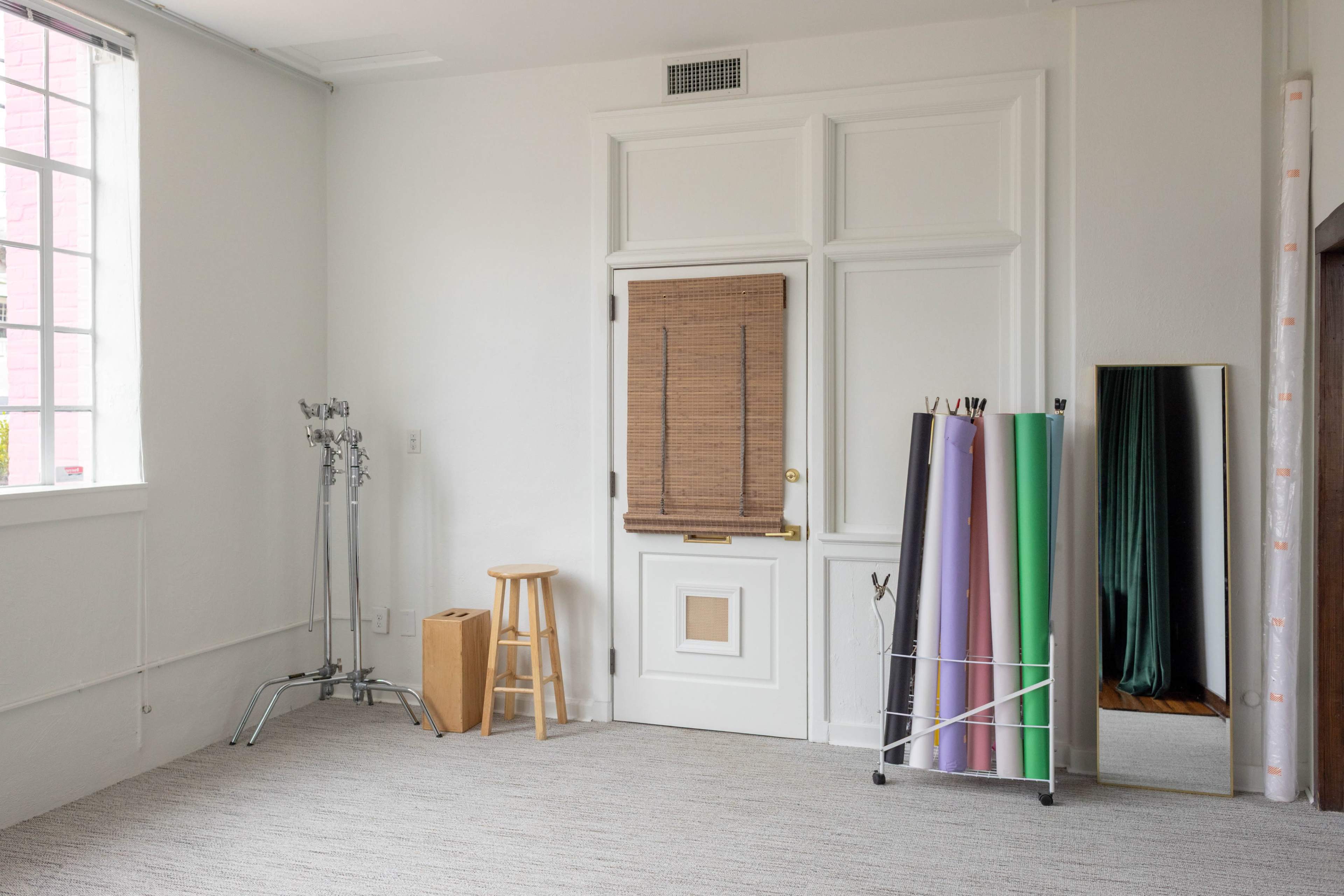 The room features a door with a wooden panel, a tall mirror, and a stand holding rolls of colored paper against a plain wall.