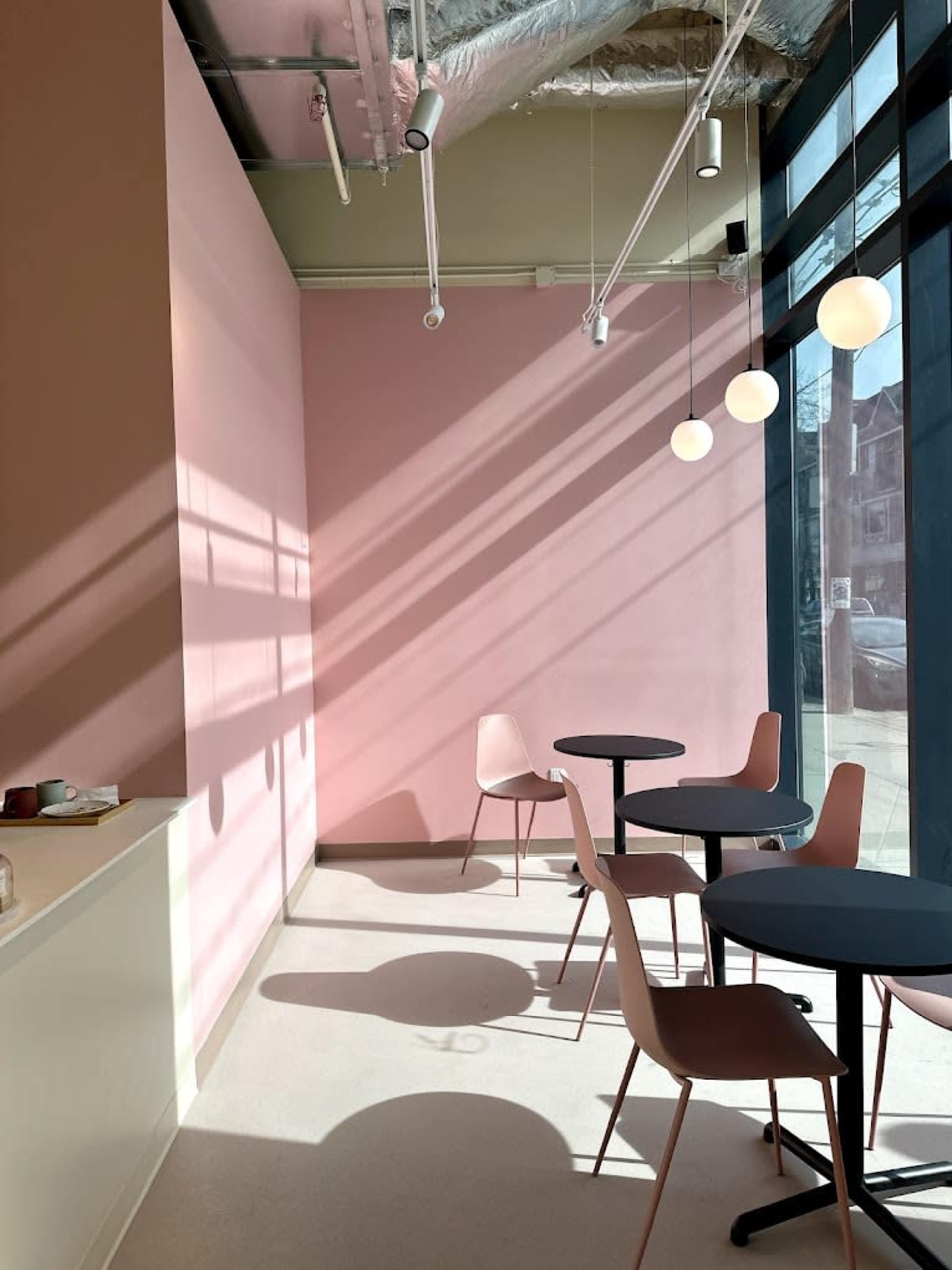 A modern café interior with pink walls, two round tables, and four pendant lights casting shadows on the floor.
