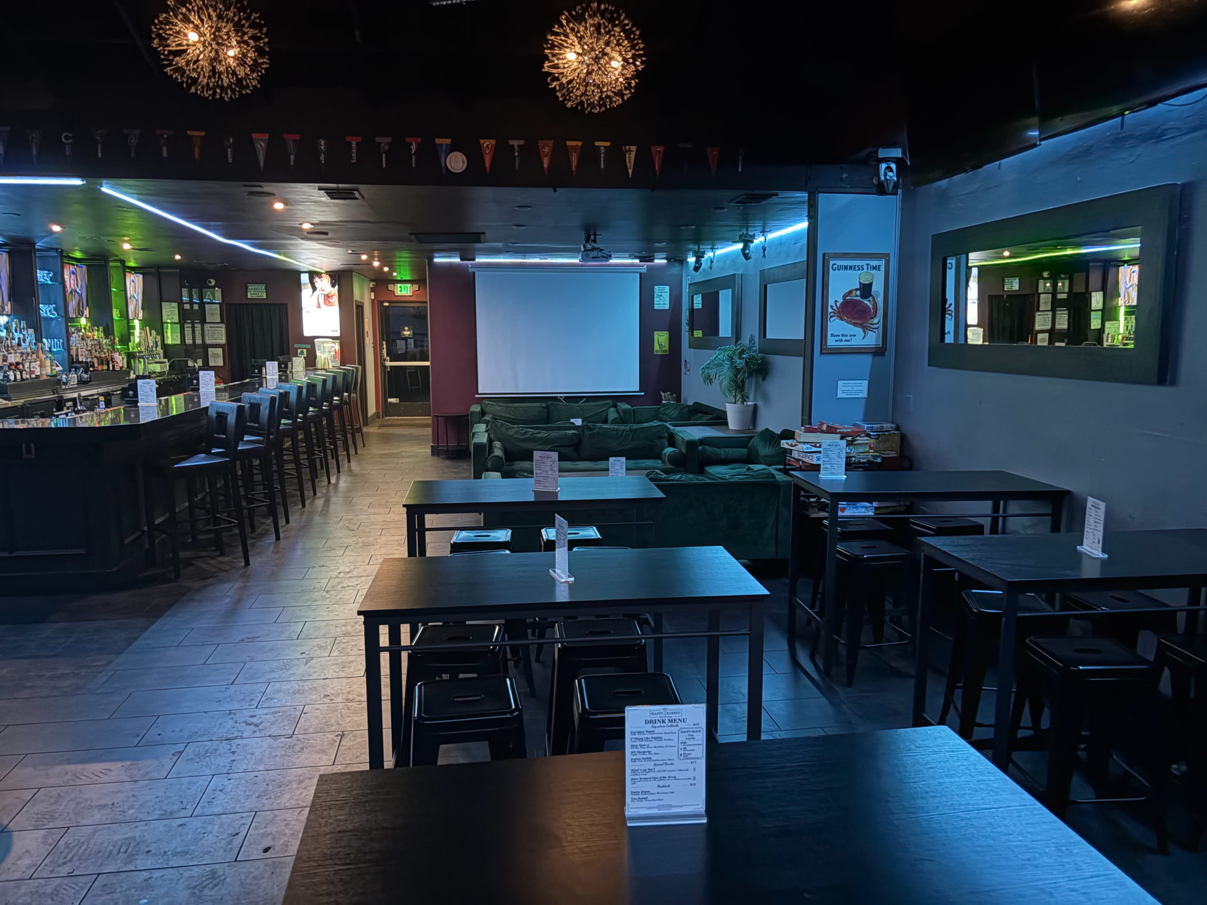 The image shows a spacious bar area with high tables and stools, a long bar counter, a projection screen at the front, and green couches along the side walls.