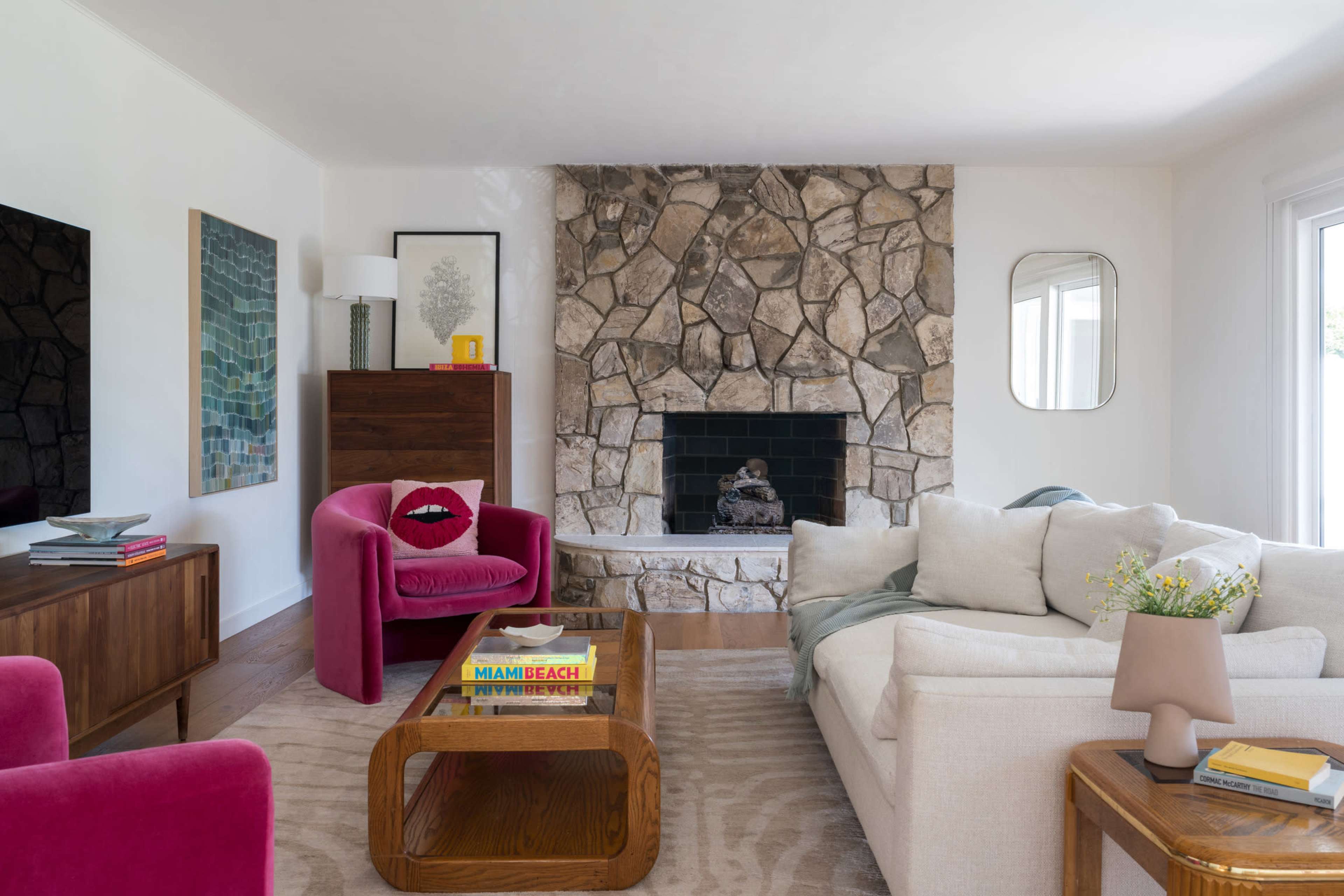 The living room features a stone fireplace, a large white sofa, a pink accent chair, and wood furniture arranged around a coffee table with books.