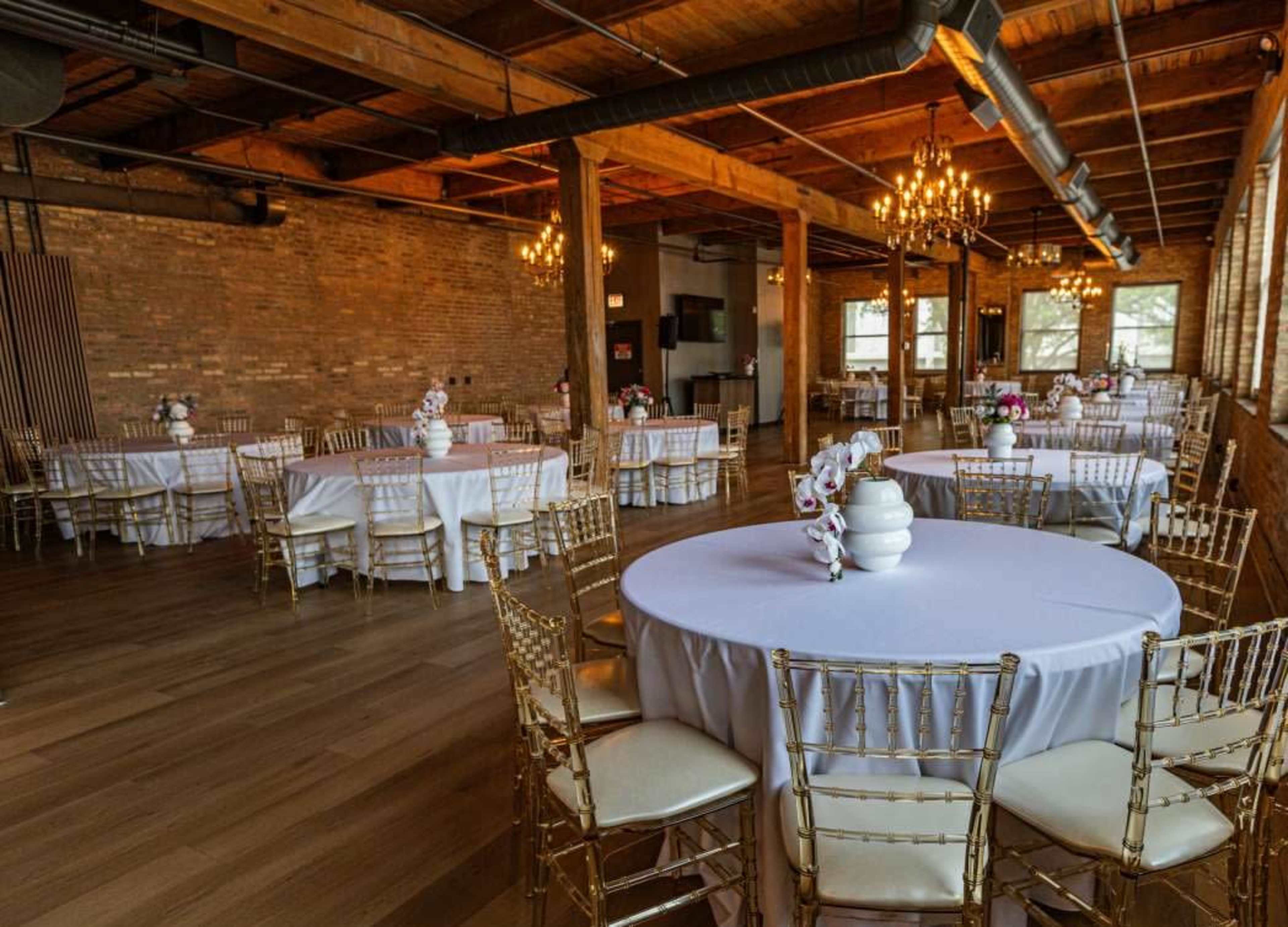 The image depicts a beautifully arranged event space with round tables covered in white tablecloths, gold chairs, and floral centerpieces, set in a room with exposed wooden beams and brick walls.