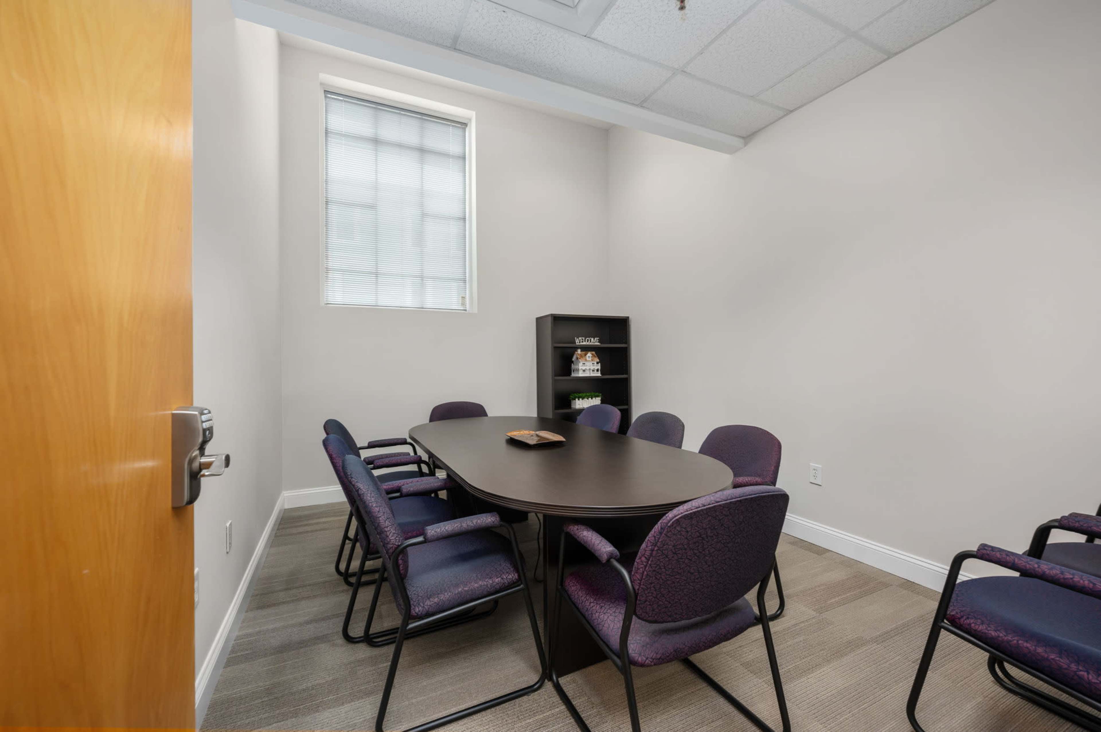 The image depicts a small conference room with a central oval table surrounded by several purple chairs, a shelf with items, and a window allowing natural light.