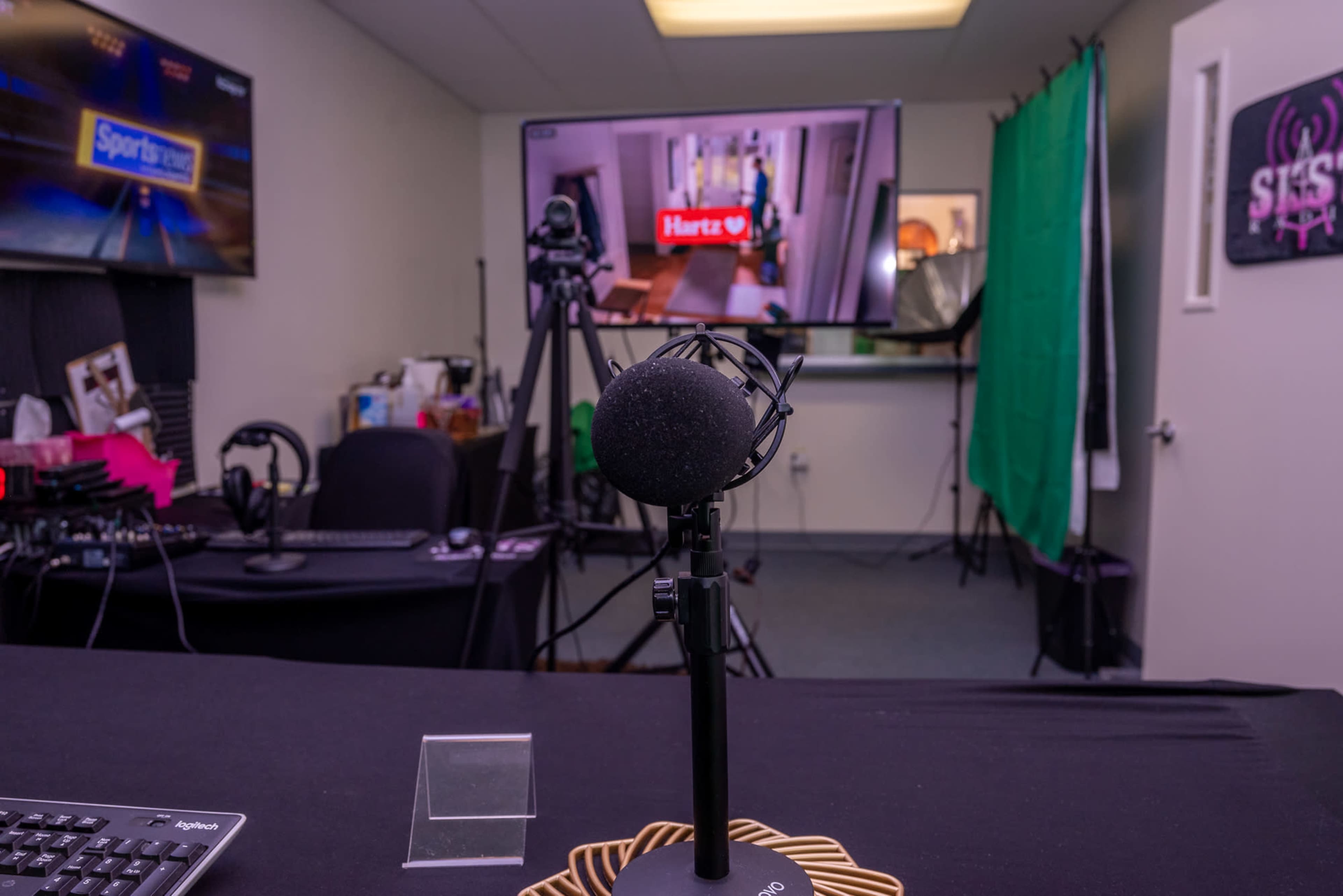 A recording studio setup with a microphone on a stand in the foreground, a keyboard, and monitors displaying various content in the background.
