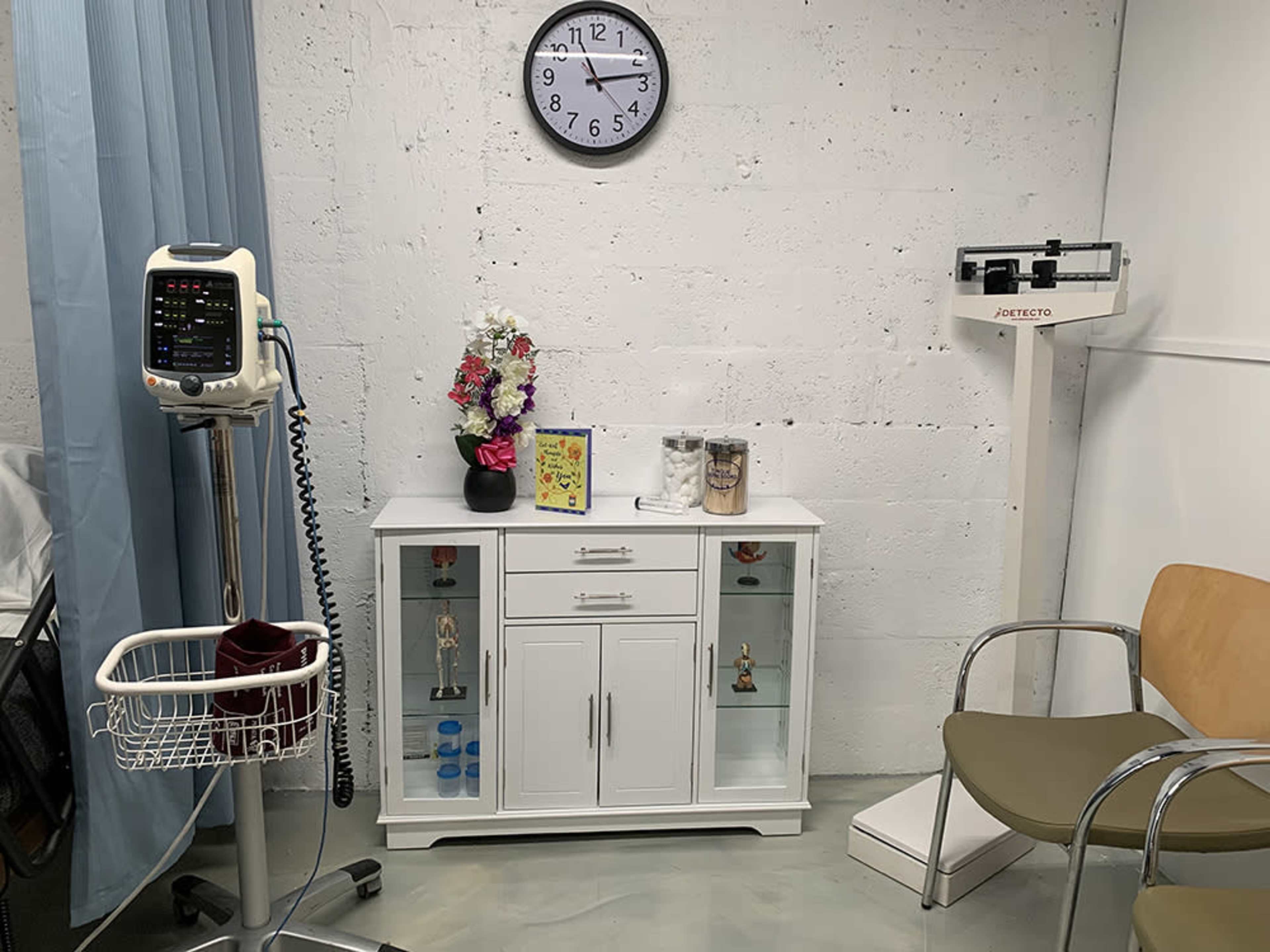 The image shows a medical examination room featuring a wall clock, a standing scale, a cabinet with decorations, and a medical monitor on a wheeled cart.