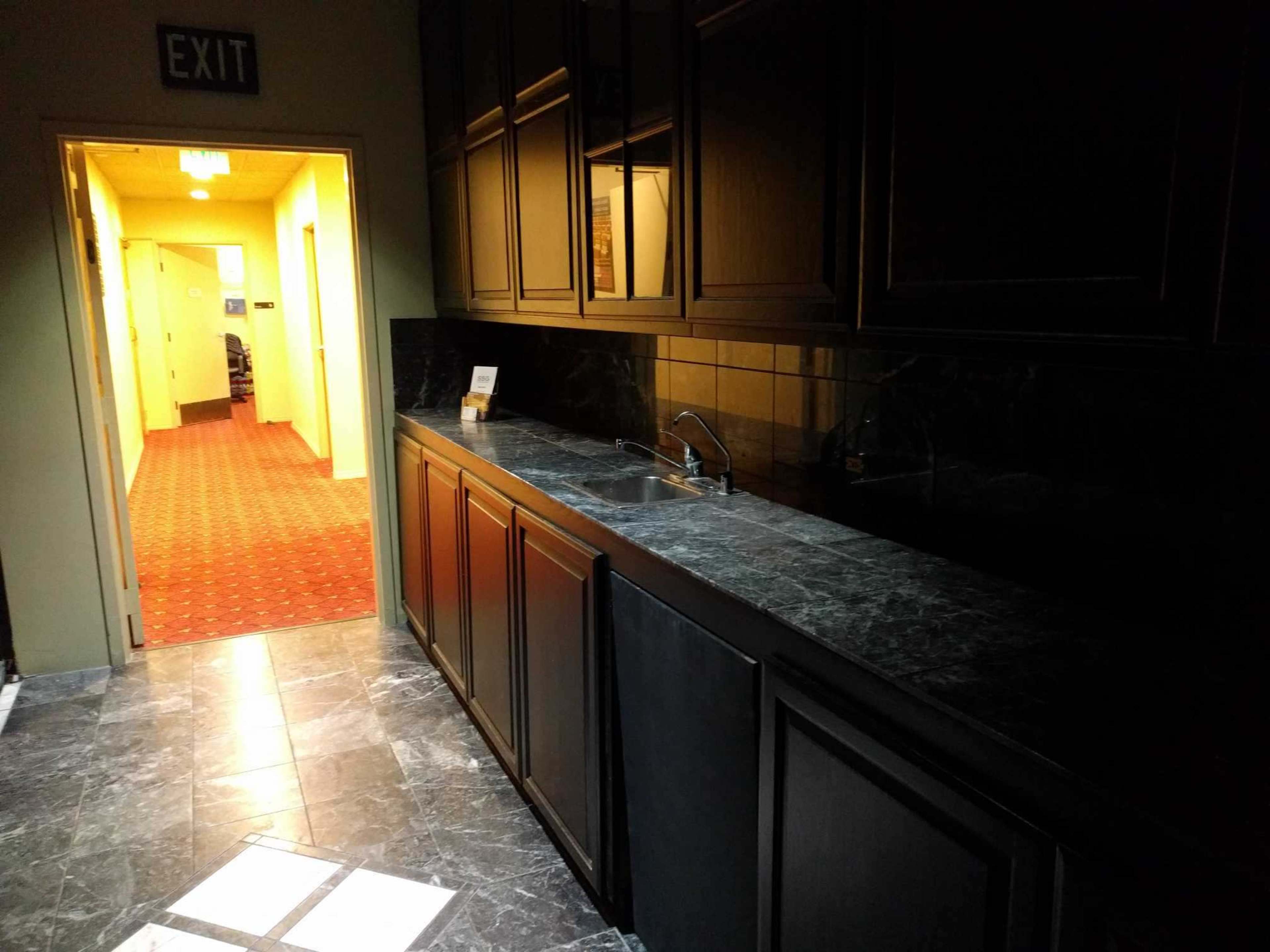 The image shows a dimly lit kitchenette with dark cabinetry and a sink, adjacent to a hallway leading to a brightly lit area.