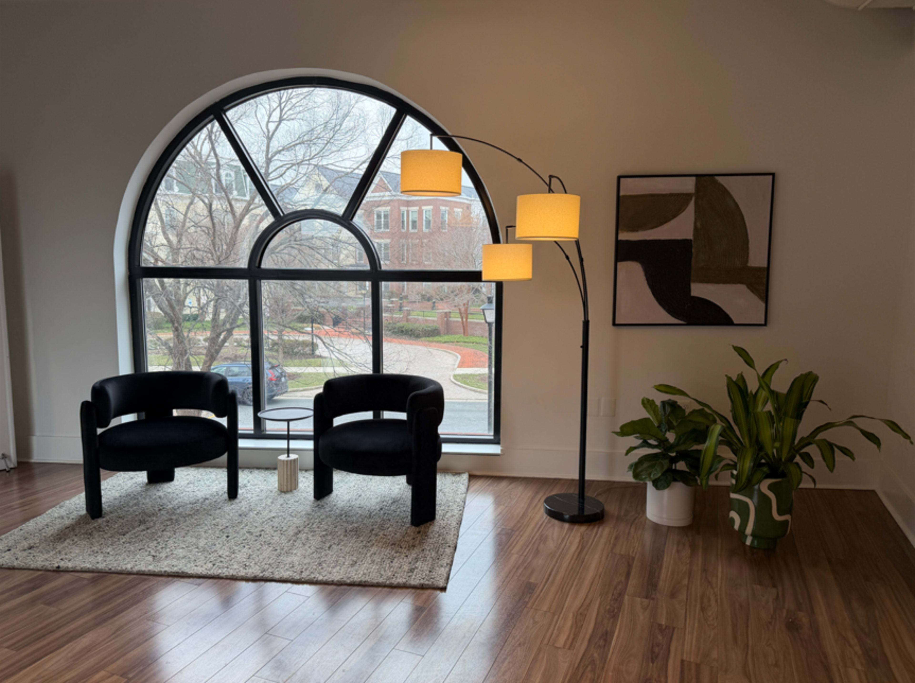 The image features two black chairs with a small table between them, located near a large arched window that overlooks a park-like setting, with a floor lamp and a potted plant nearby.