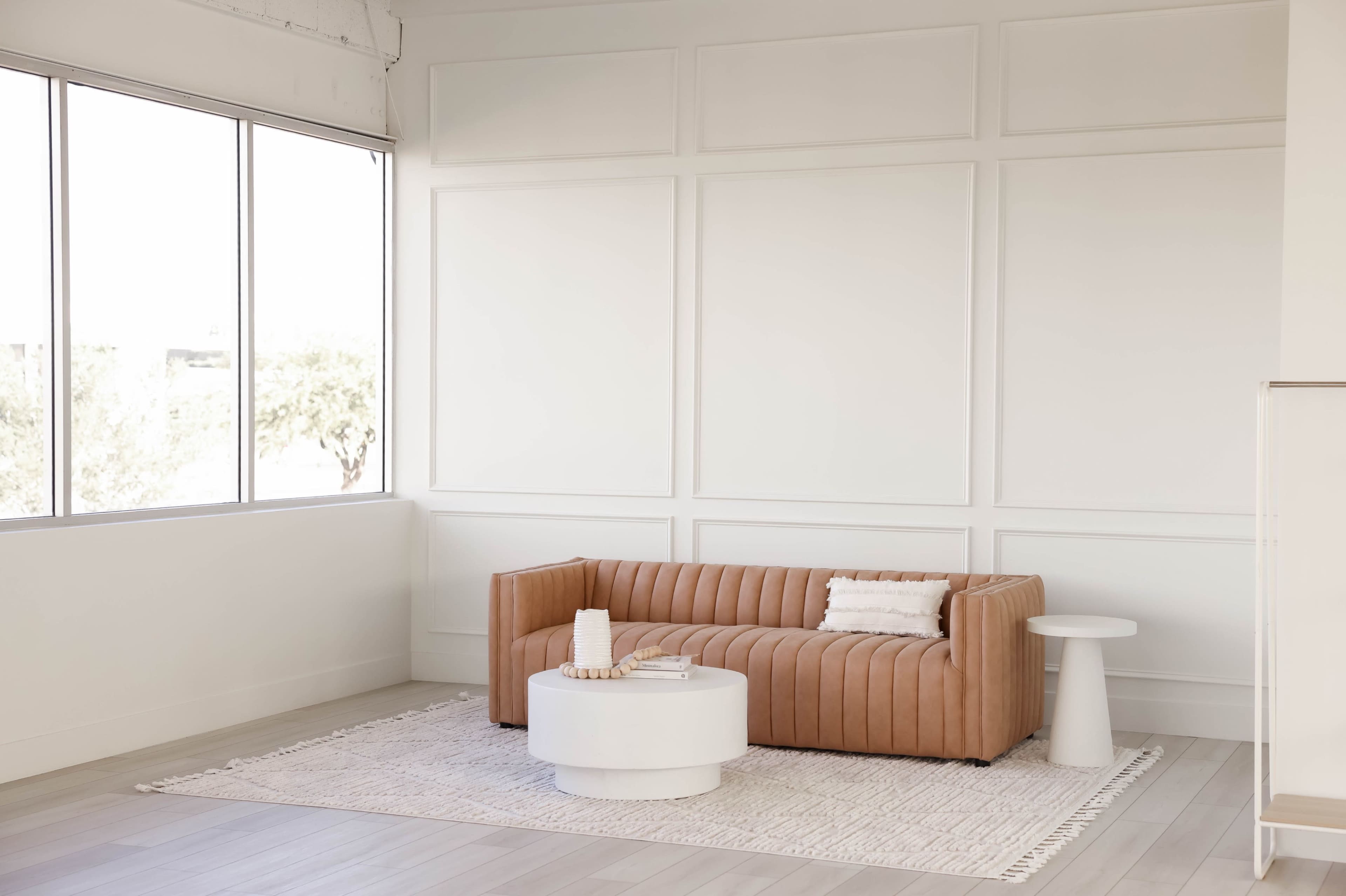 A tan velvet sofa is positioned next to a round white coffee table on a light-colored rug in a room with large windows and minimalist decor.